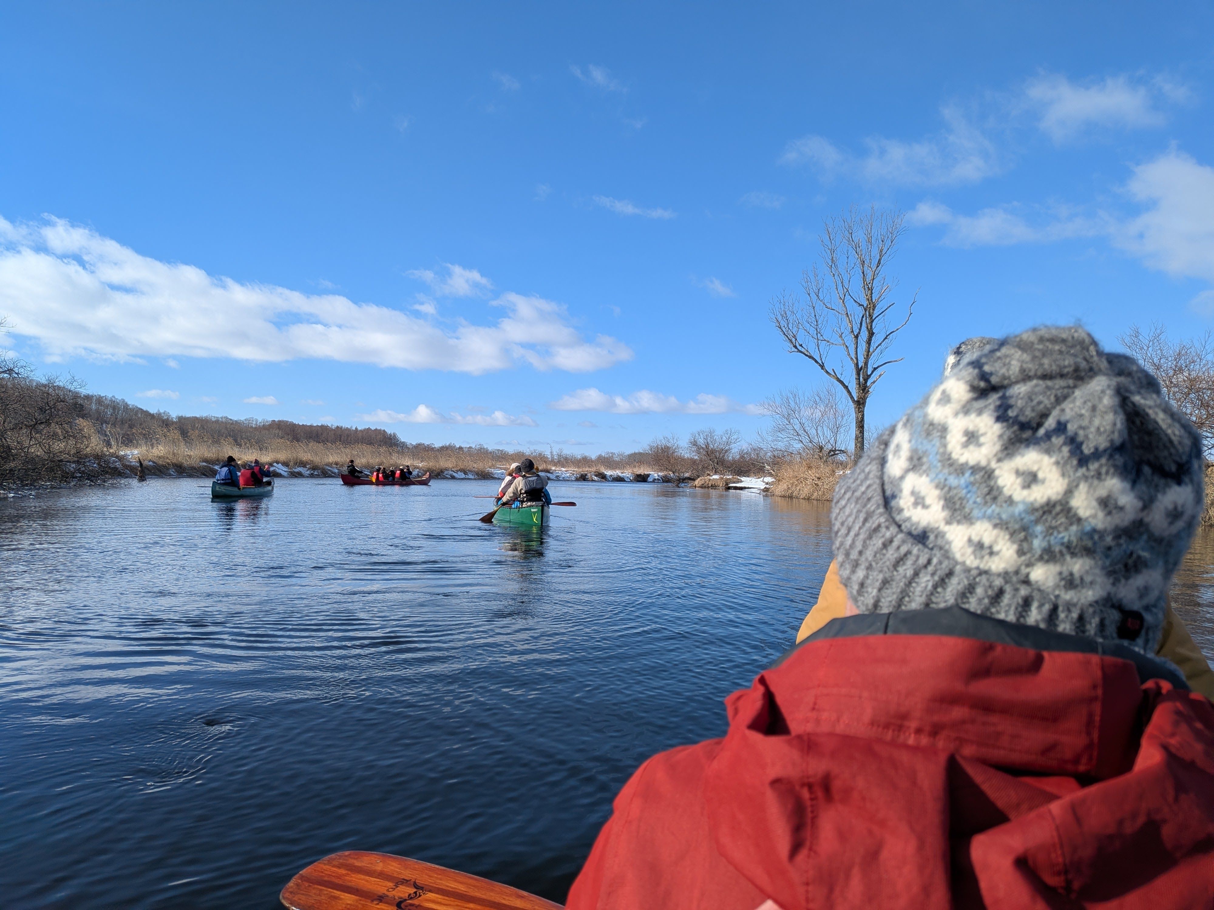 A photo taken from a canoe on the water's surface. The photographer is behind someone wearing a woolly hat. In the distance, three other canoes paddle ahead further downstream.