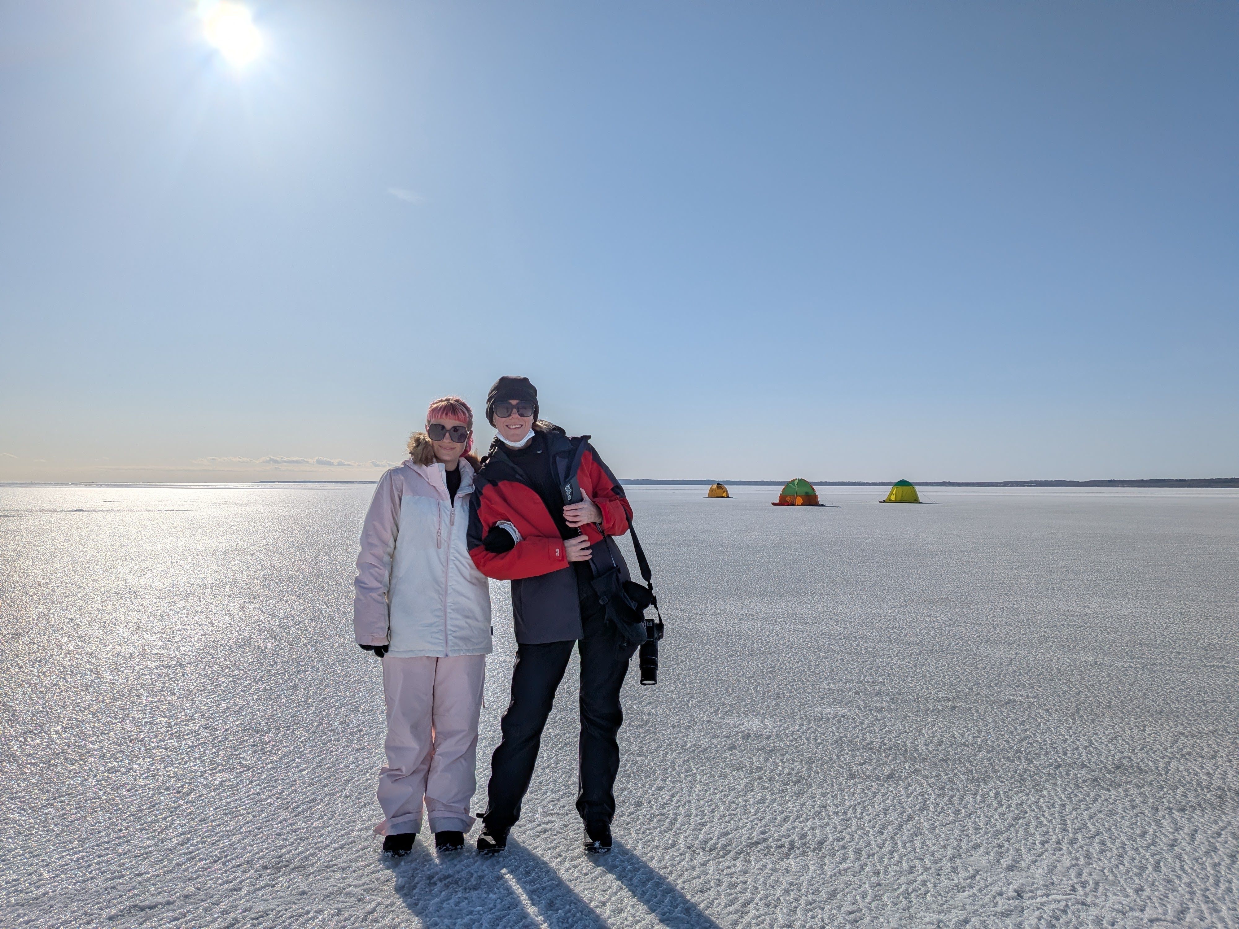 Two women smile at the camera, stood on the vast ice expanse on Notsuke Peninsula. It is a sunny day with not a cloud in the sky. In the distance, small tents where people are ice fishing are visible.