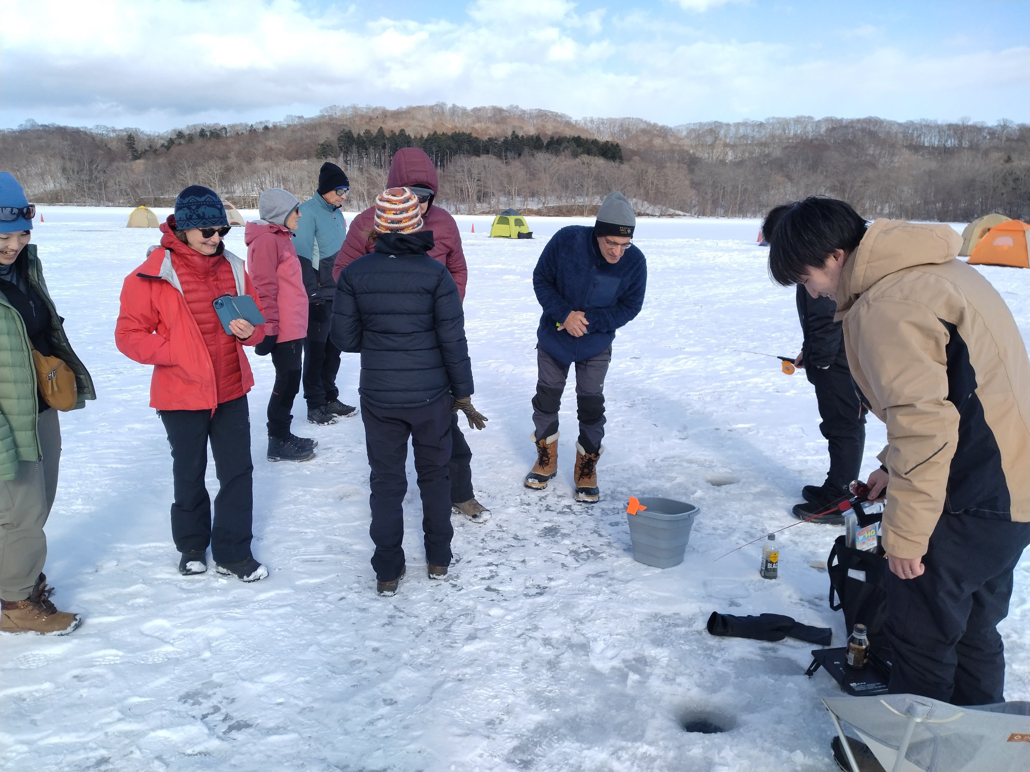 A group of people gather around a man ice fishing through holes on a frozen lake. There is a bucket in which he has his catch and one in the group is peeping inside.