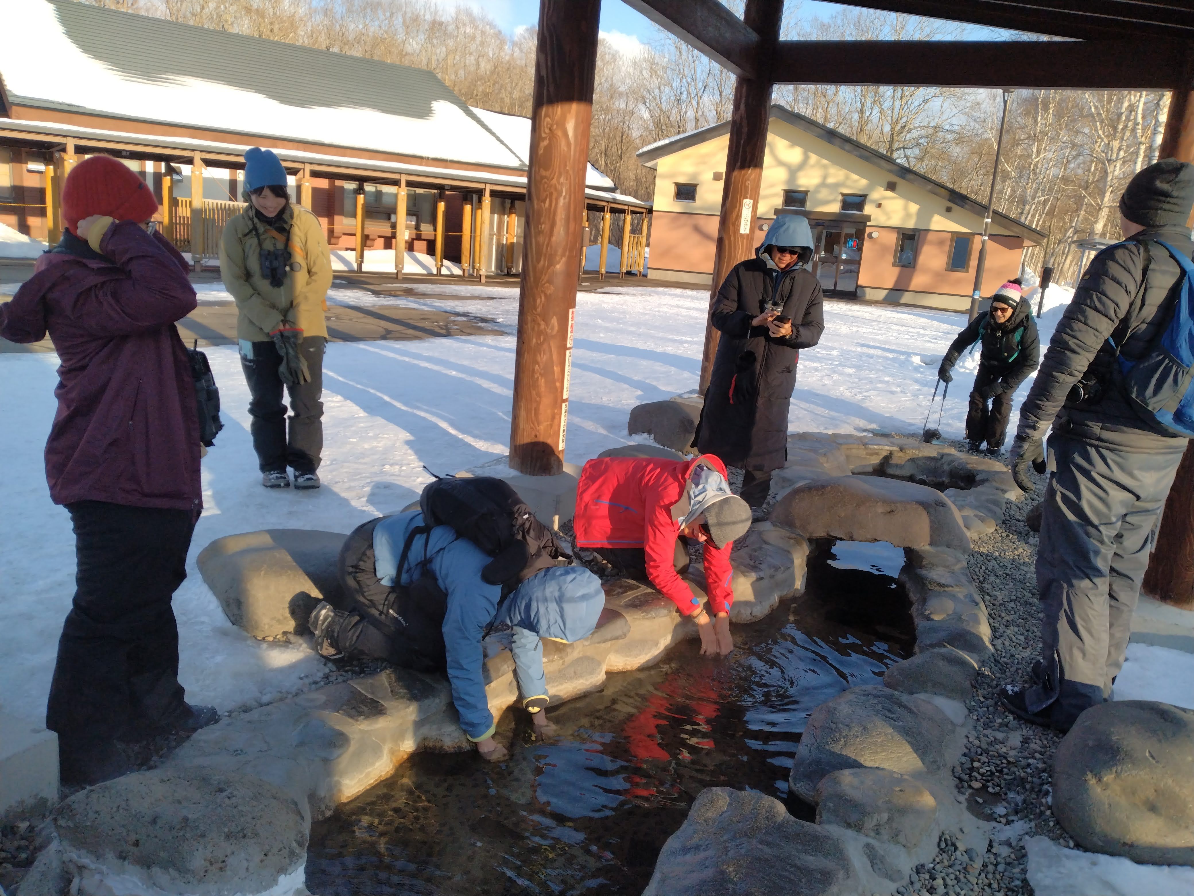 A group of people in winter garb stand around a natural hot spring pond for feet, known as an "ashiyu" in Japanese. Some of them are putting their hands into the water to warm their hands on this cold winter's day.