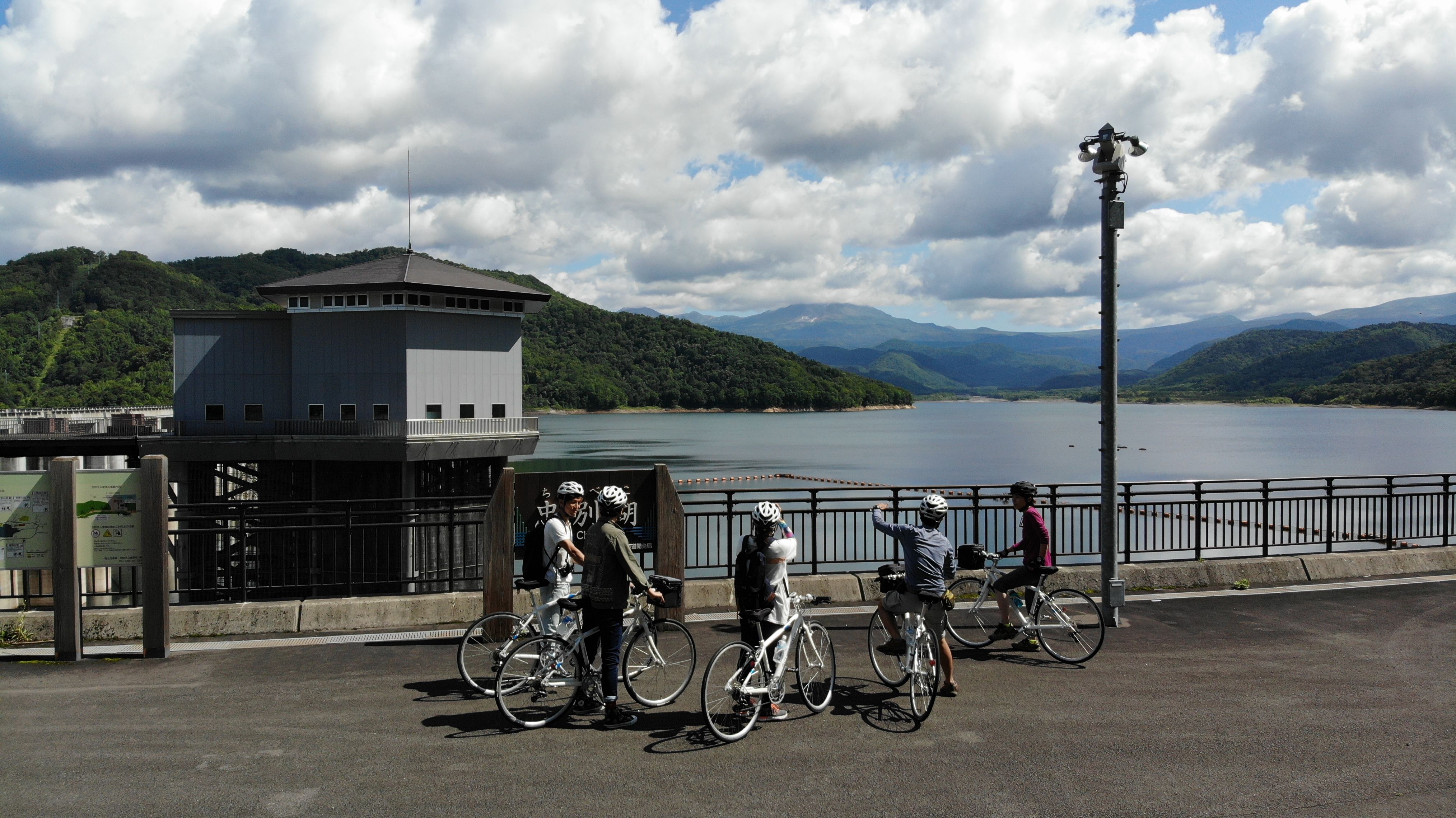 A group of five cyclists are dismounted next to their bikes on a dam wall, looking out over Lake Chubetsu in Higashikawa, Hokkaido. The lake is a dam lake. It is a partially cloudy day.