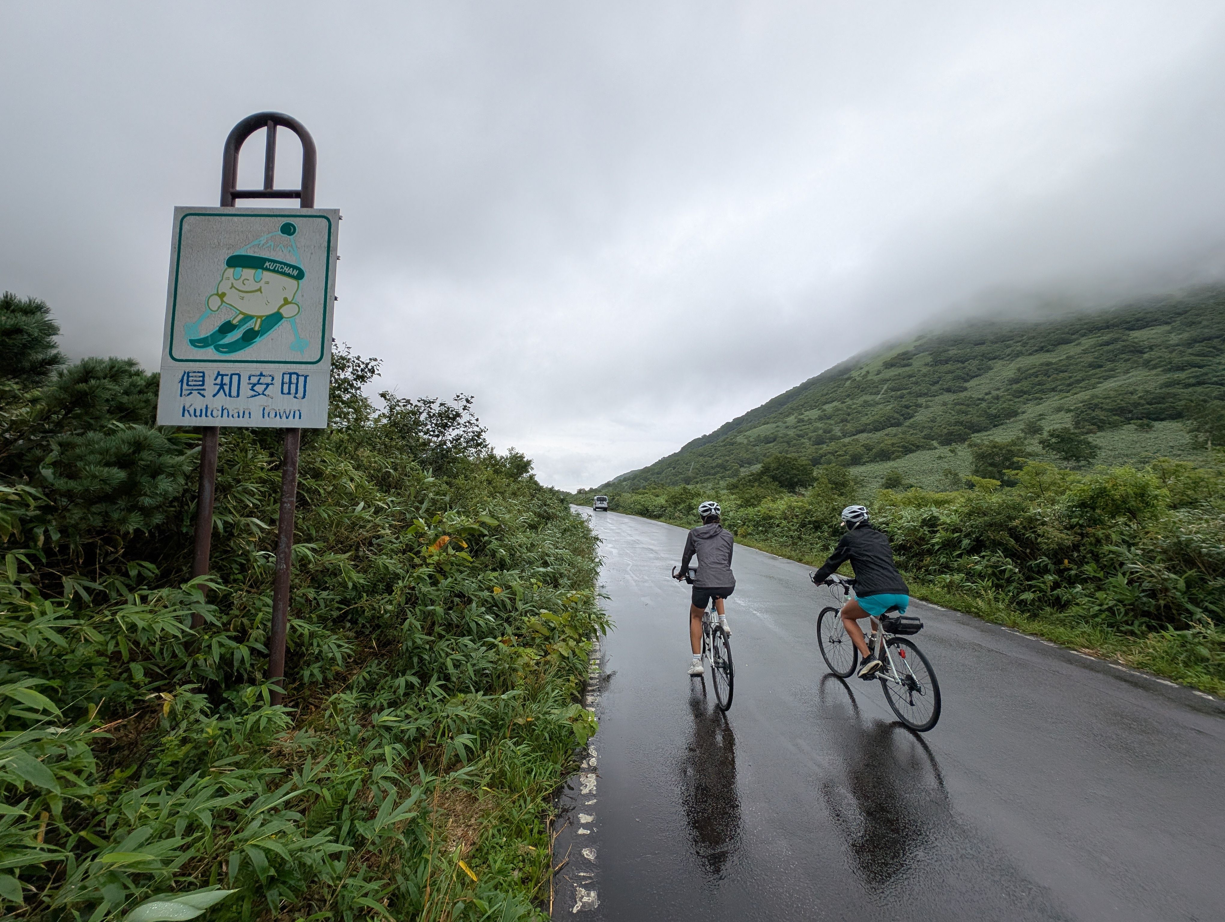 Two cyclists pass a town demarcation sign. The sign reads "Kutchan Town" and depicts the town mascot skiing. Rain has recently fallen (or is falling) and the road is wet. The clouds are low and partially obscure a mountain on the right side of the image.