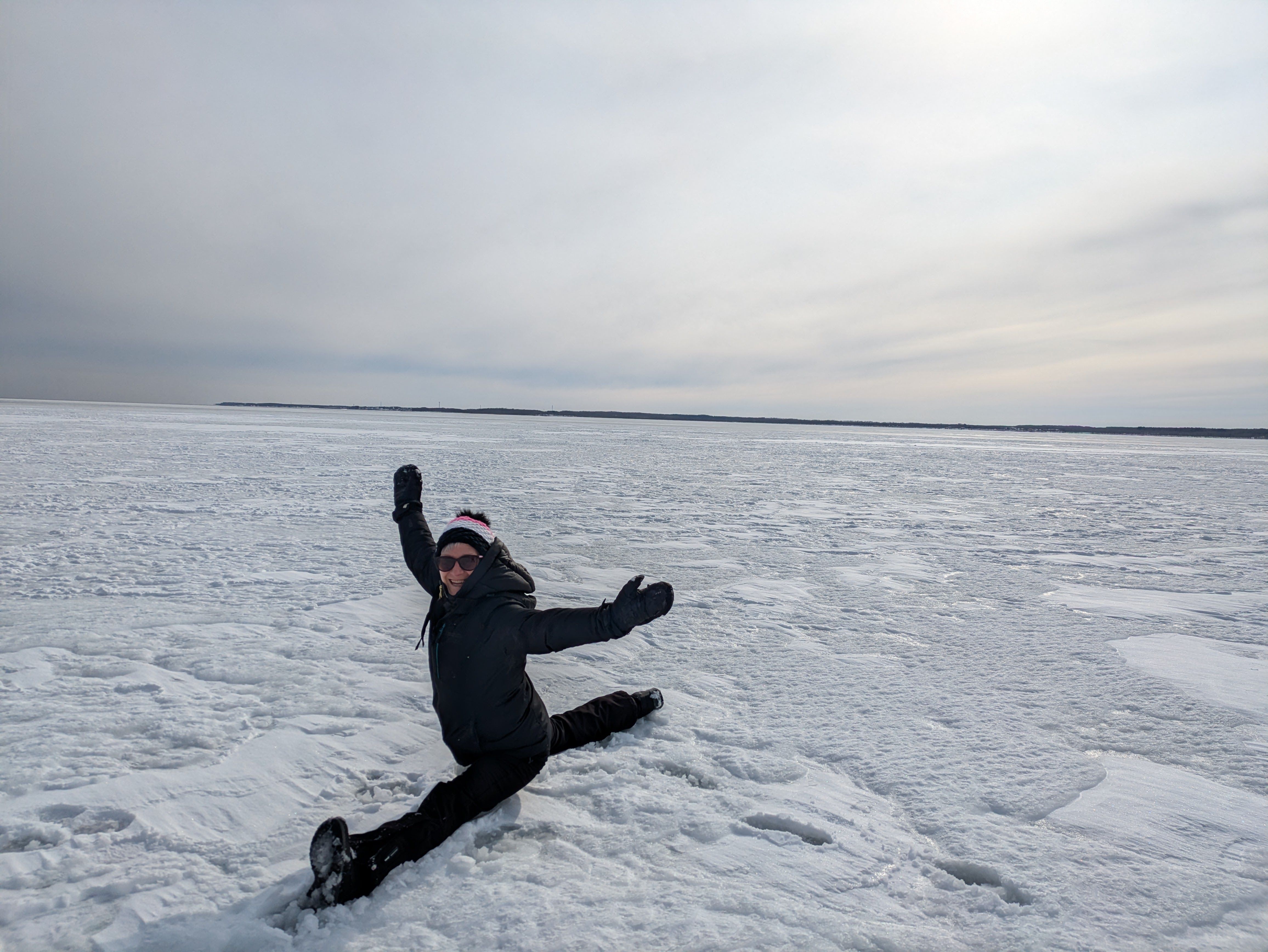 A woman on an ice field does the splits and waves her arms in the air, smiling at the camera.