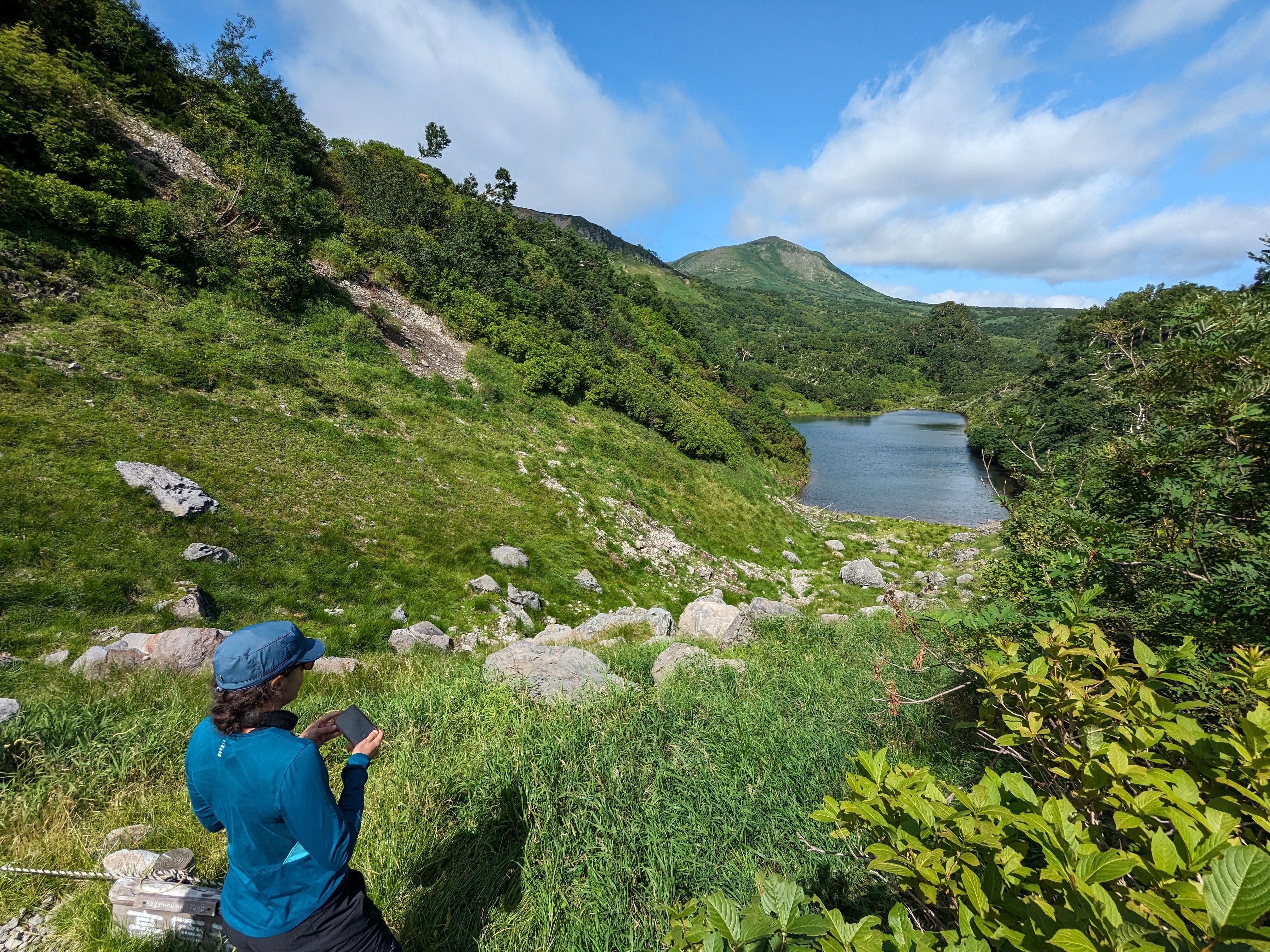 A lady stands in front of a mountain landscape, a pond nestled in a valley below.