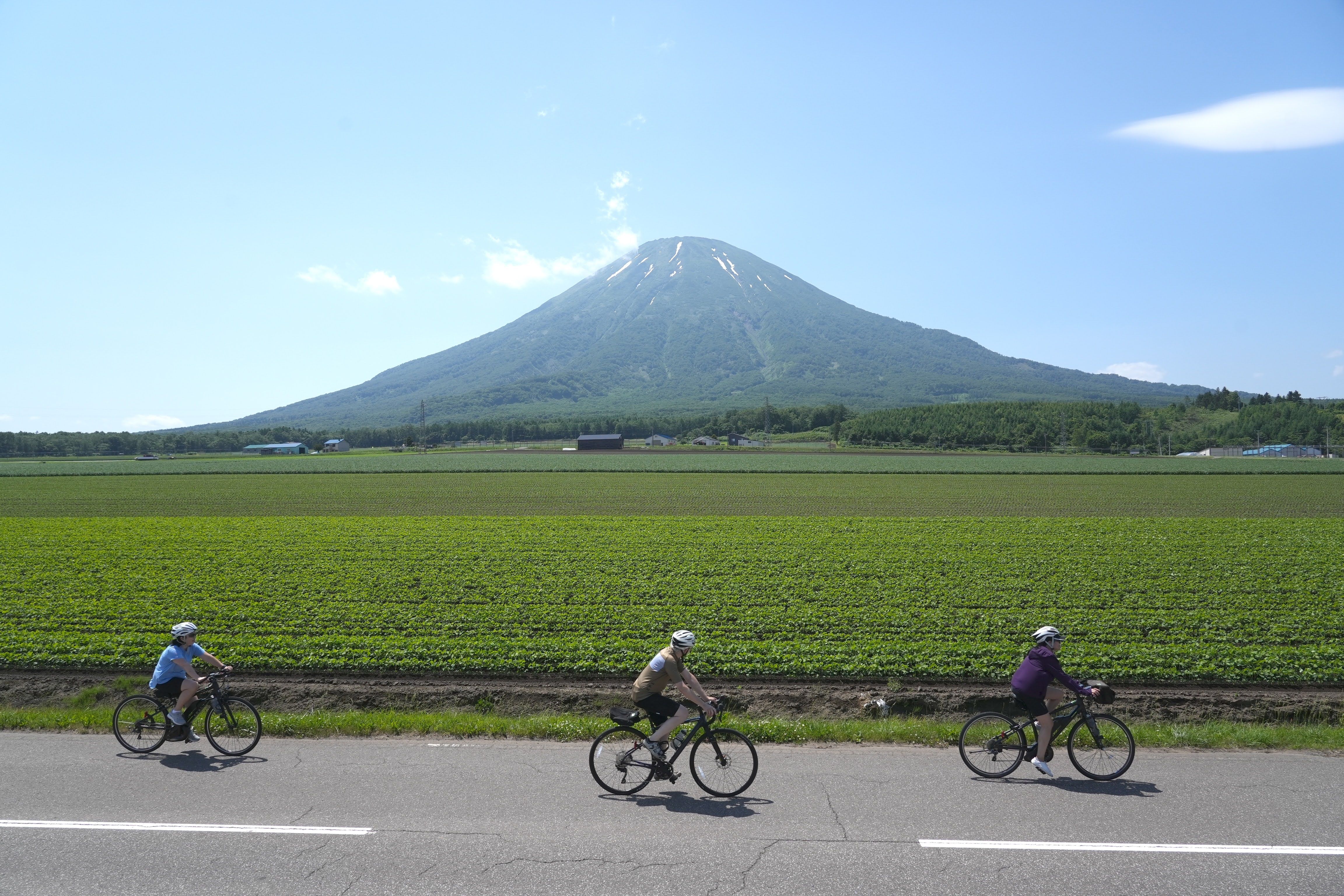 Three cyclists ride past Mt. Yotei in Hokkaido. It is a very sunny day and the mountain is clearly visible. Some patches of snow remain visible at its summit. Farmland surrounds the cyclists.
