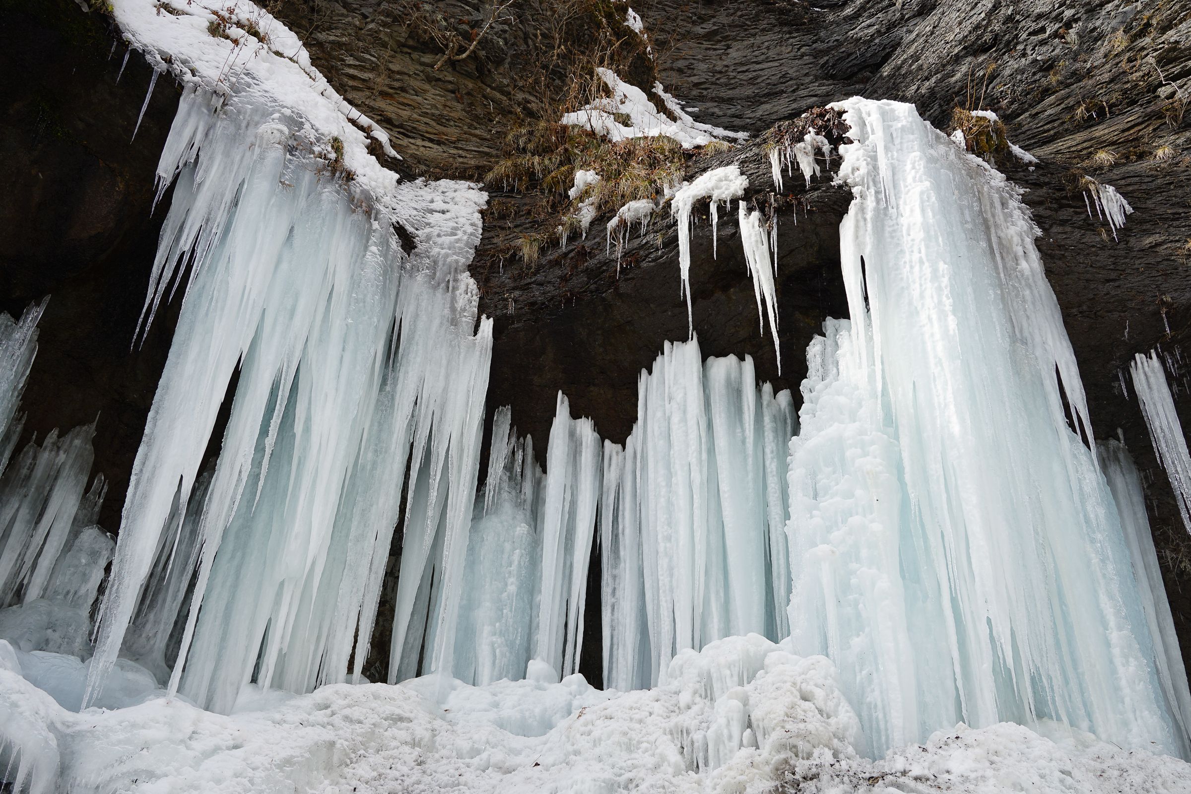 A dramatic Hokkaido winter scene featuring massive, jagged icicles and thick frozen ice pillars hanging from a dark, craggy rock face. The ice glows with a pale blue tint against the white snow and dark grey stone.