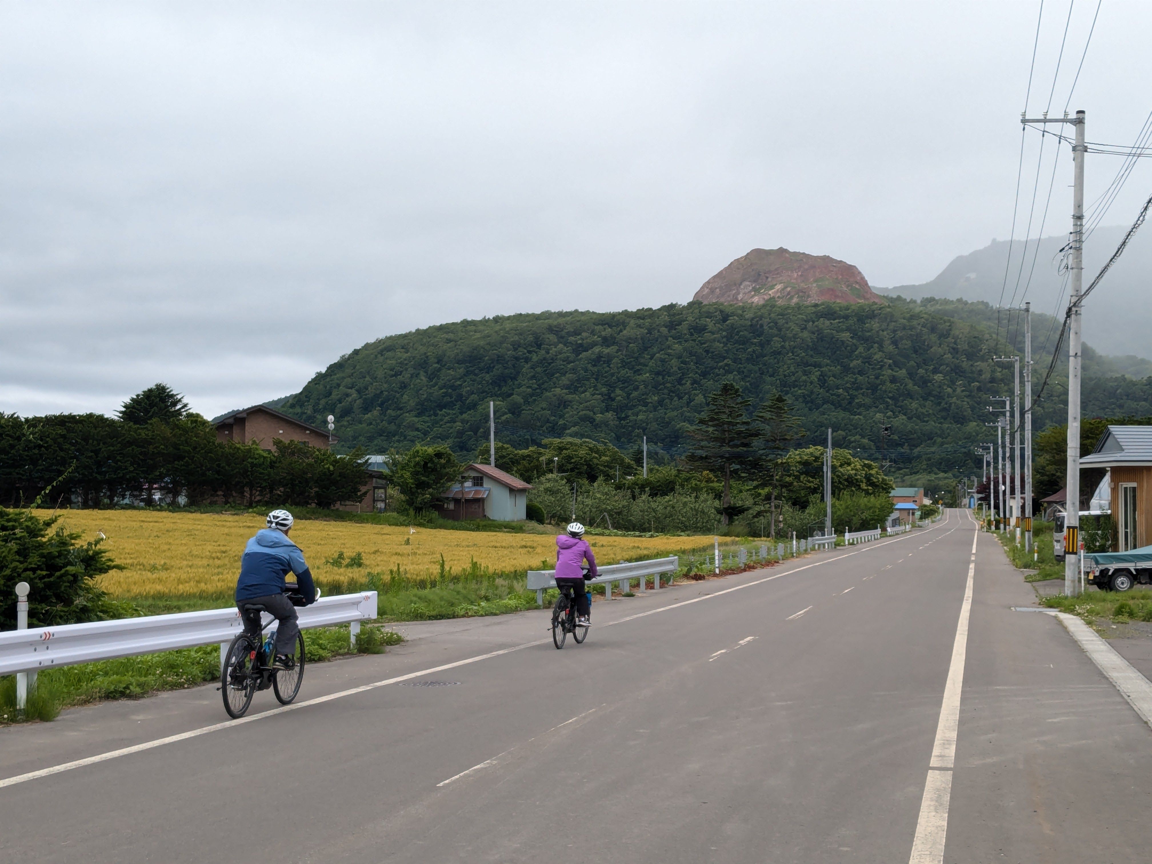 Two cyclists ride along a road approaching Showa Shinzan, a lava dome in Hokkaido. The top of the lava dome juts out of a forested mountain in the distance.