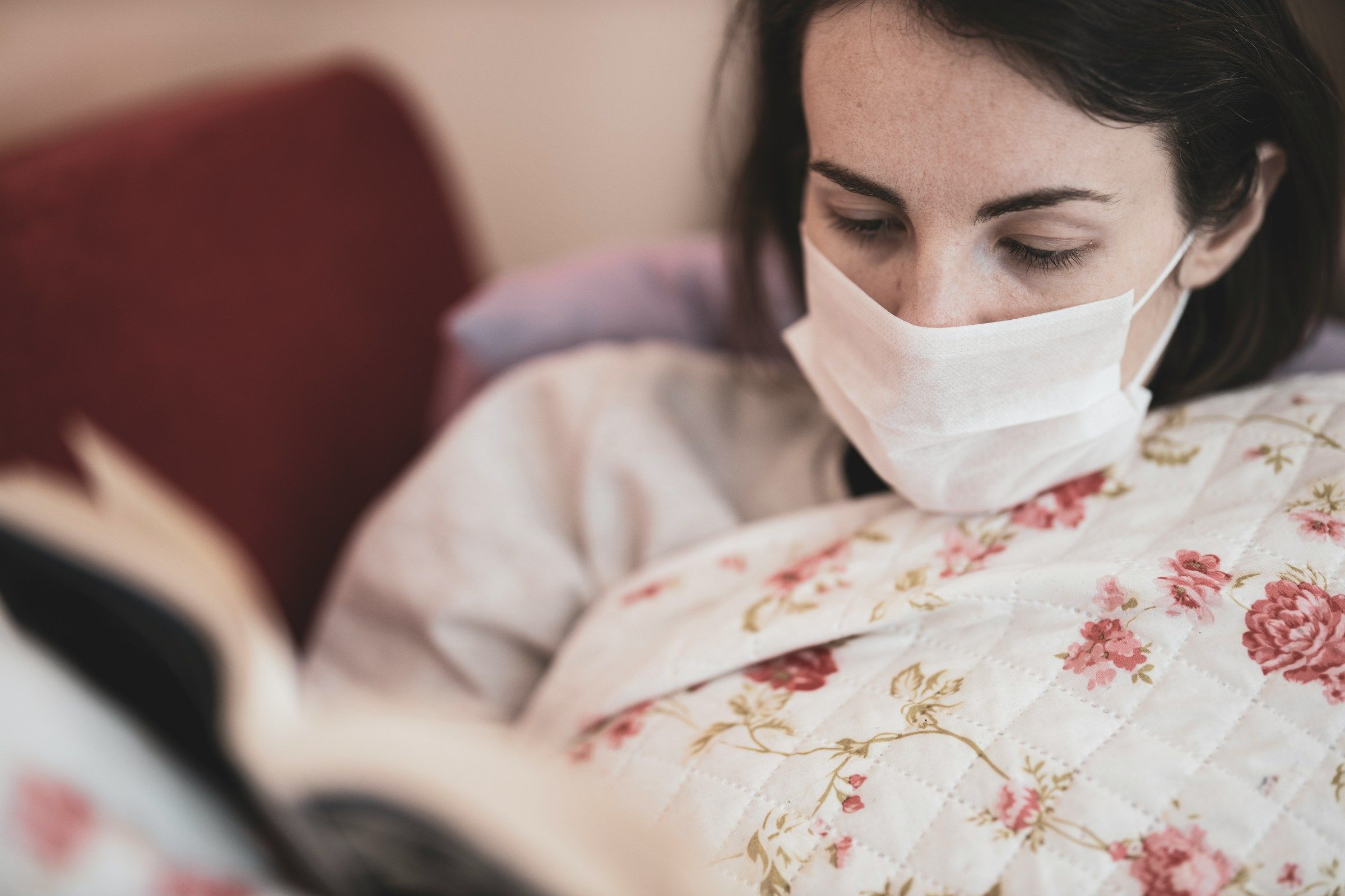 A woman wearing a face mask rests under a floral quilt while reading a book.