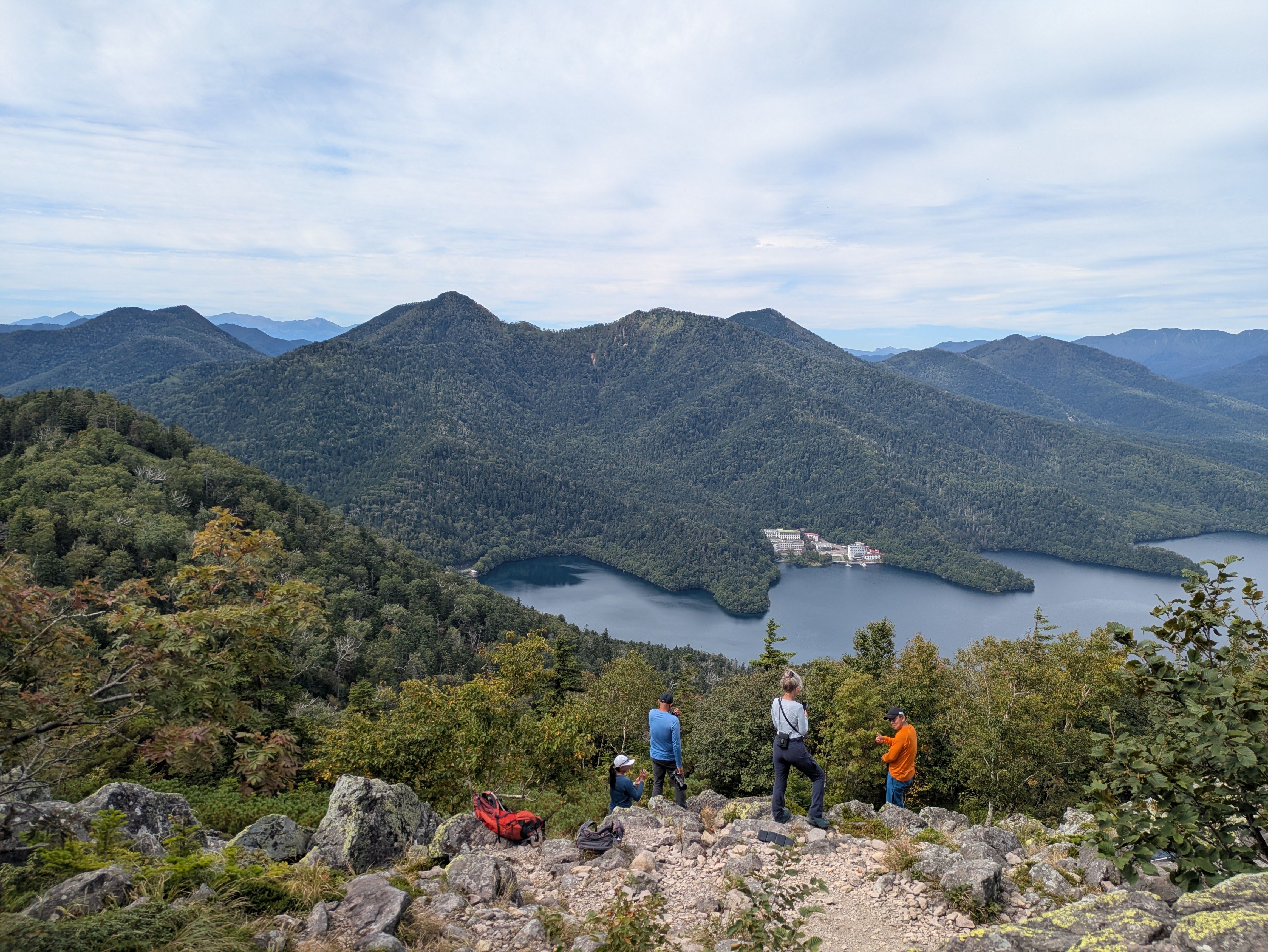 A group of hikers stands at the rocky summit of Mt. Hakuun in Hokkaido. Below them, Lake Shikaribetsu is visible in a valley. It is a slightly cloudy day.