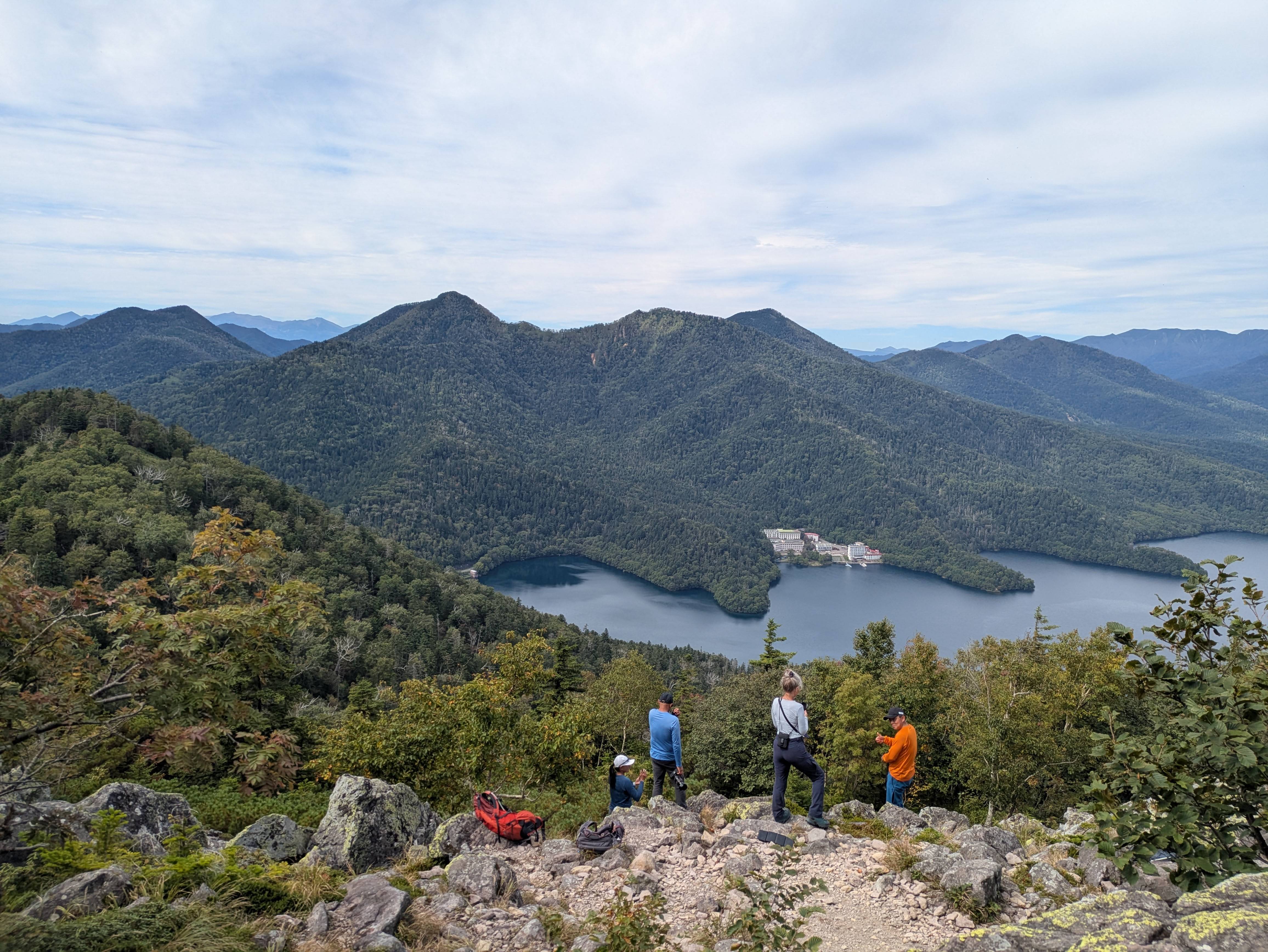 A group of hikers stands at the rocky summit of Mt. Hakuun in Hokkaido. Below them, Lake Shikaribetsu is visible in a valley. It is a slightly cloudy day.