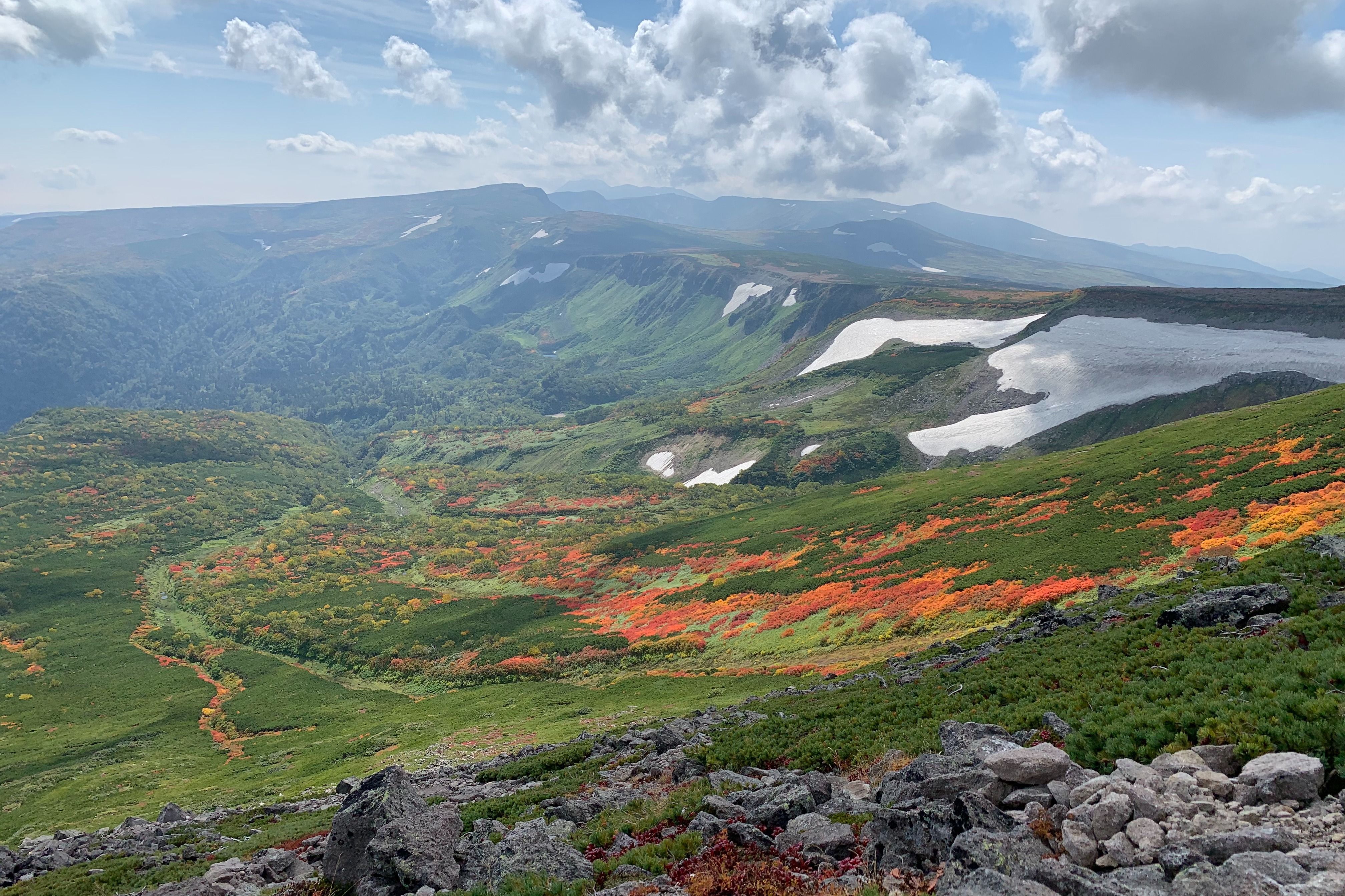 A view from the slope of Mt. Midori showing striking autumn colors slashing down through green expanses of pine. Snow at a slightly higher elevation offsets this contrast of color with a wintery white.
