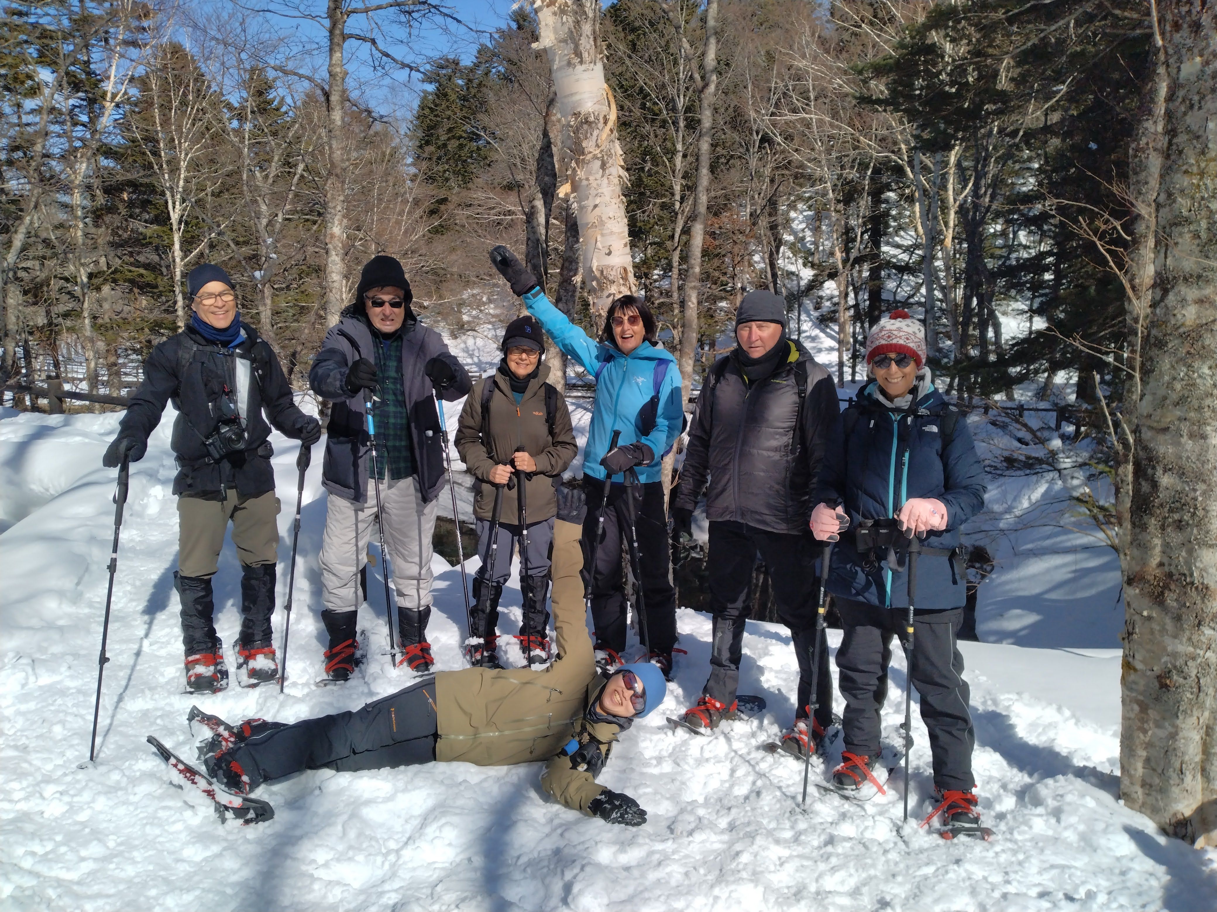 A group of people in winter gear and snowshoes in a forest smile, pose and wave for the camera. It is a very sunny day.