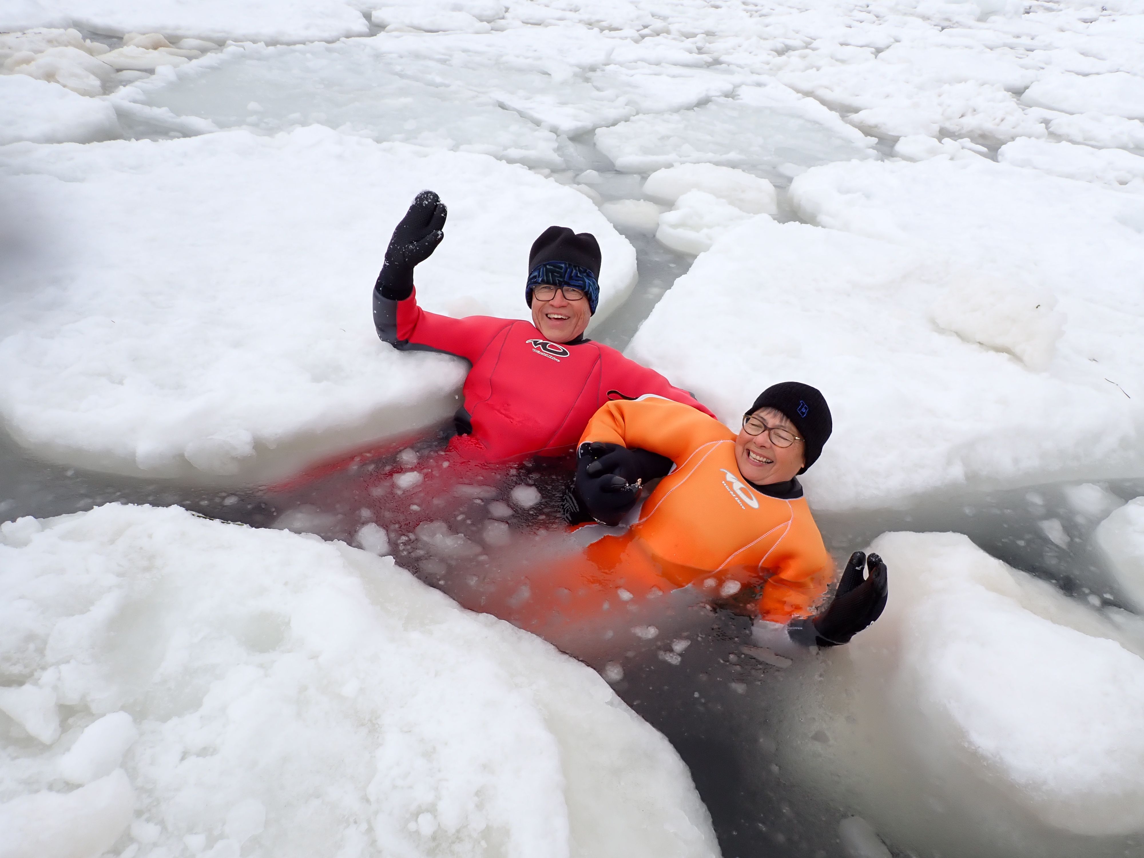 Two people wearing drysuits smile and wave at the camera as they float in the ocean between drift ice floes. They don't look cold, thankfully, thanks to their drysuits!