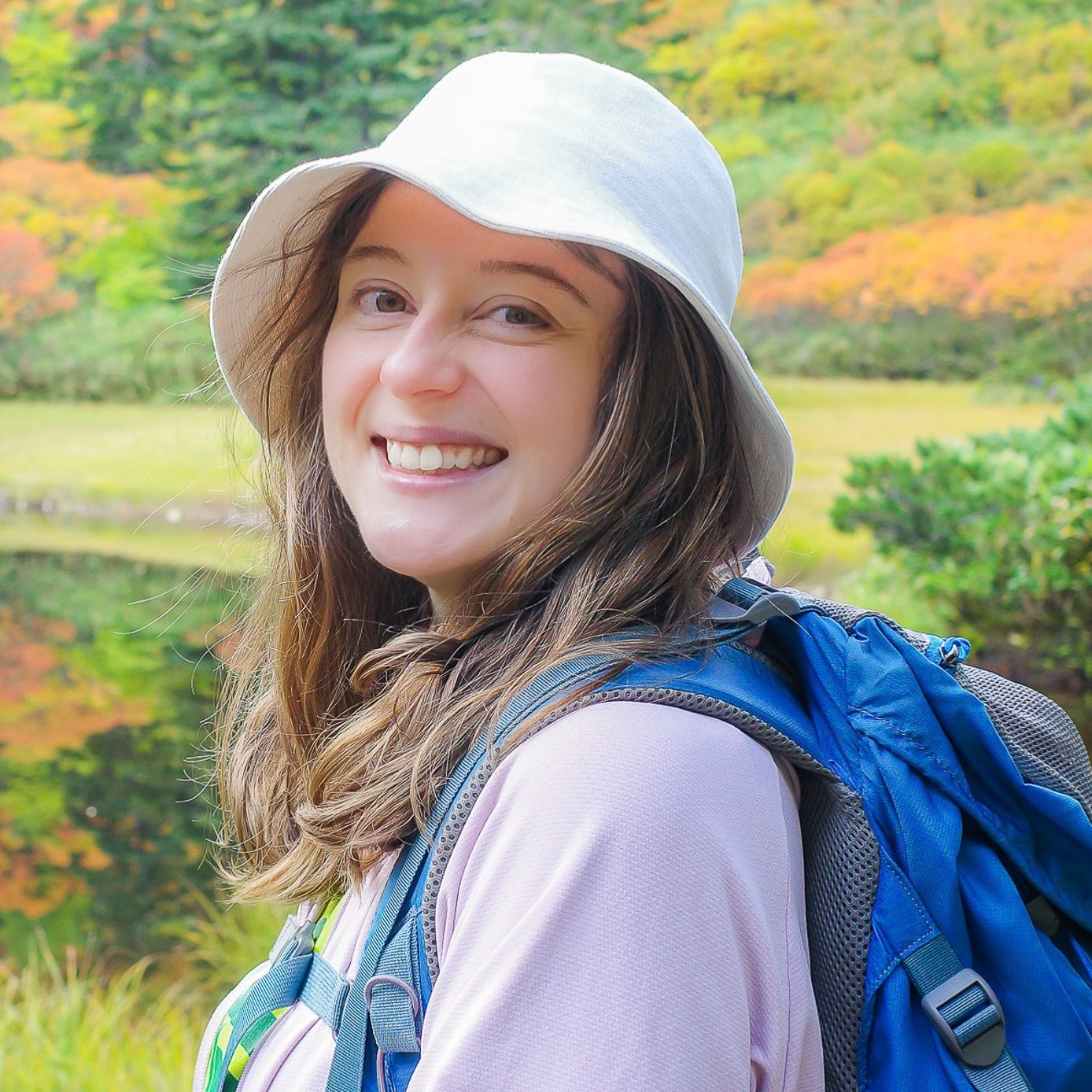 A female hiker wearing a cute white hat beams into the camera. In the background the first autumn colours in the Daisetsuzan are reflected in a still pond at Kogen Onsen.