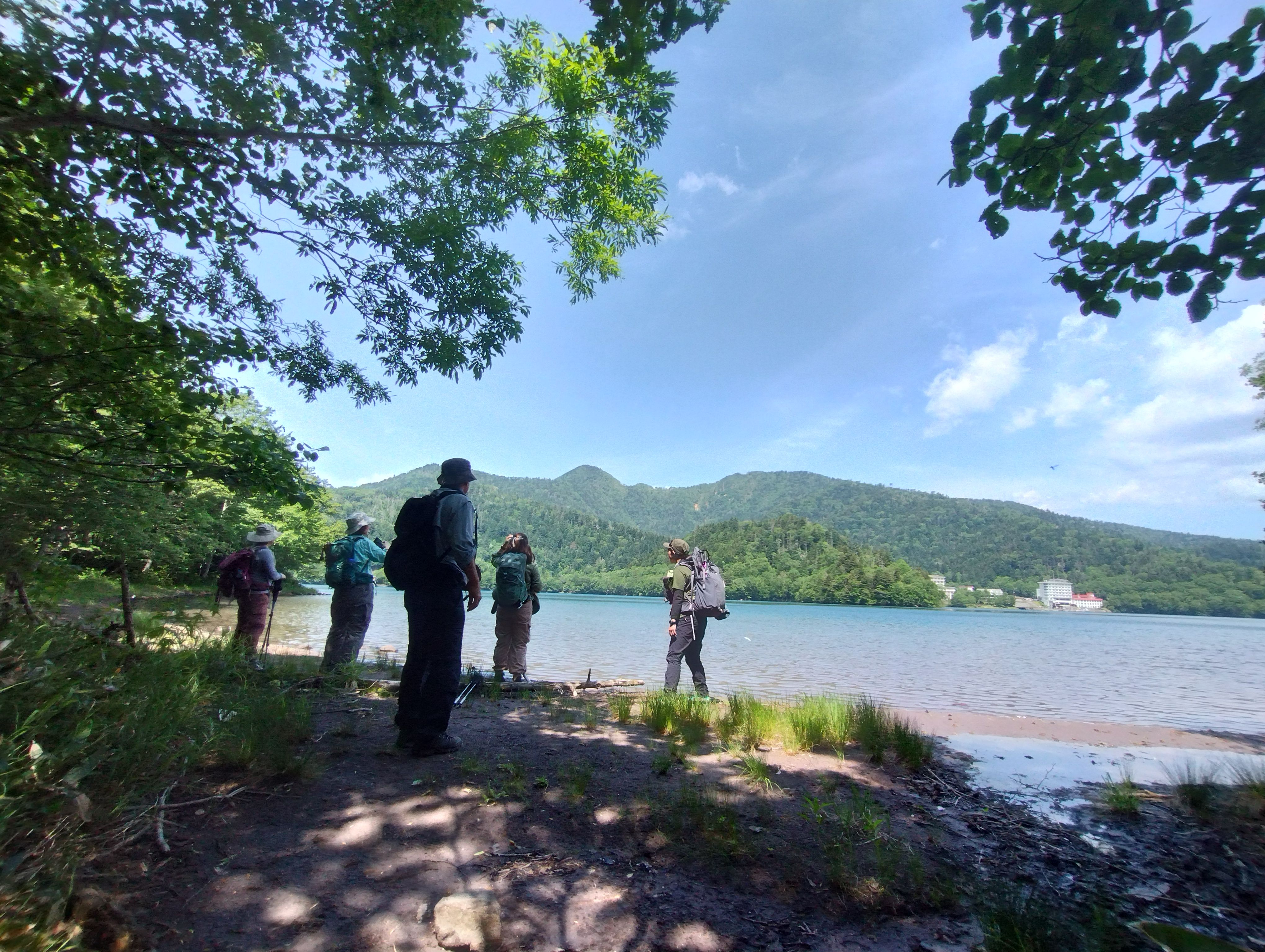 A group of hikers gather on the shore of Lake Shikaribetsu in Hokkaido. It is a sunny day with blue skies overhead.