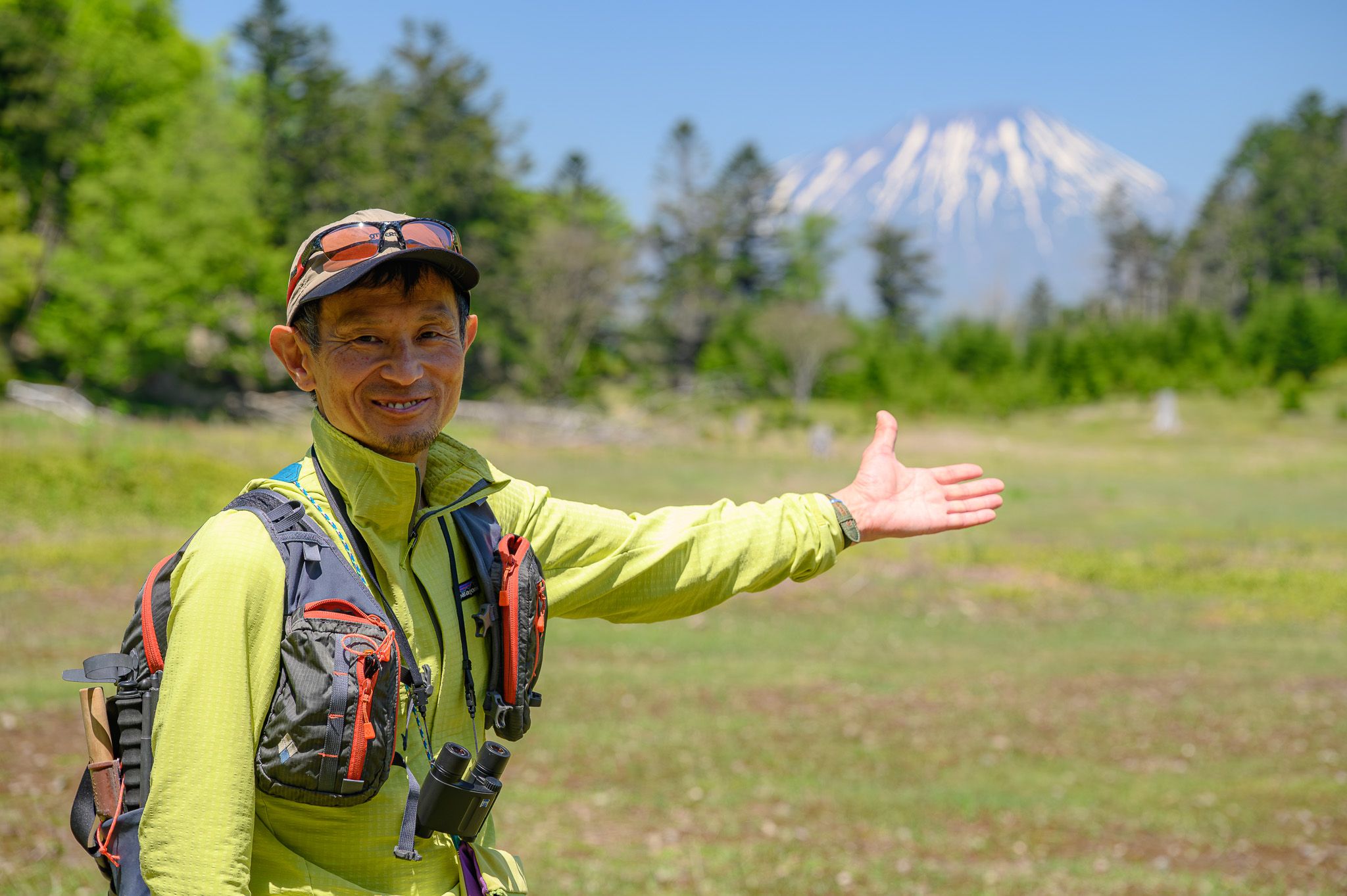Adventure Hokkaido guide Gen poses at Daiheigen on Nakajima Island, Lake Toya. In the background, the summit of Mt. Yotei rises in the gap between green trees and blue sky.