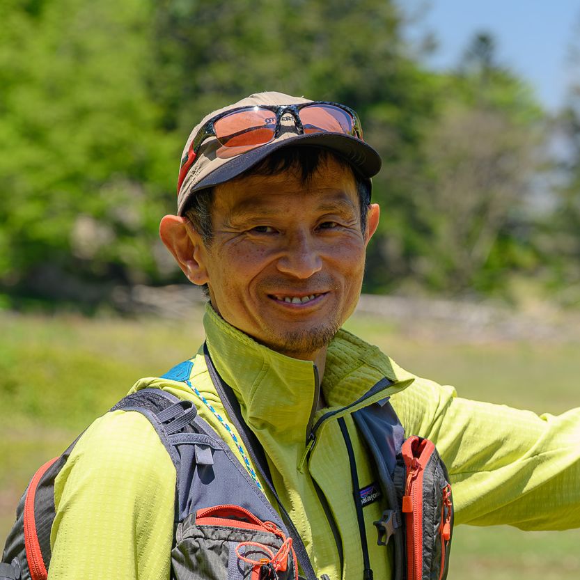 Adventure Hokkaido guide Gen poses at Daiheigen on Nakajima Island, Lake Toya. In the background, the summit of Mt. Yotei rises in the gap between green trees and blue sky.
