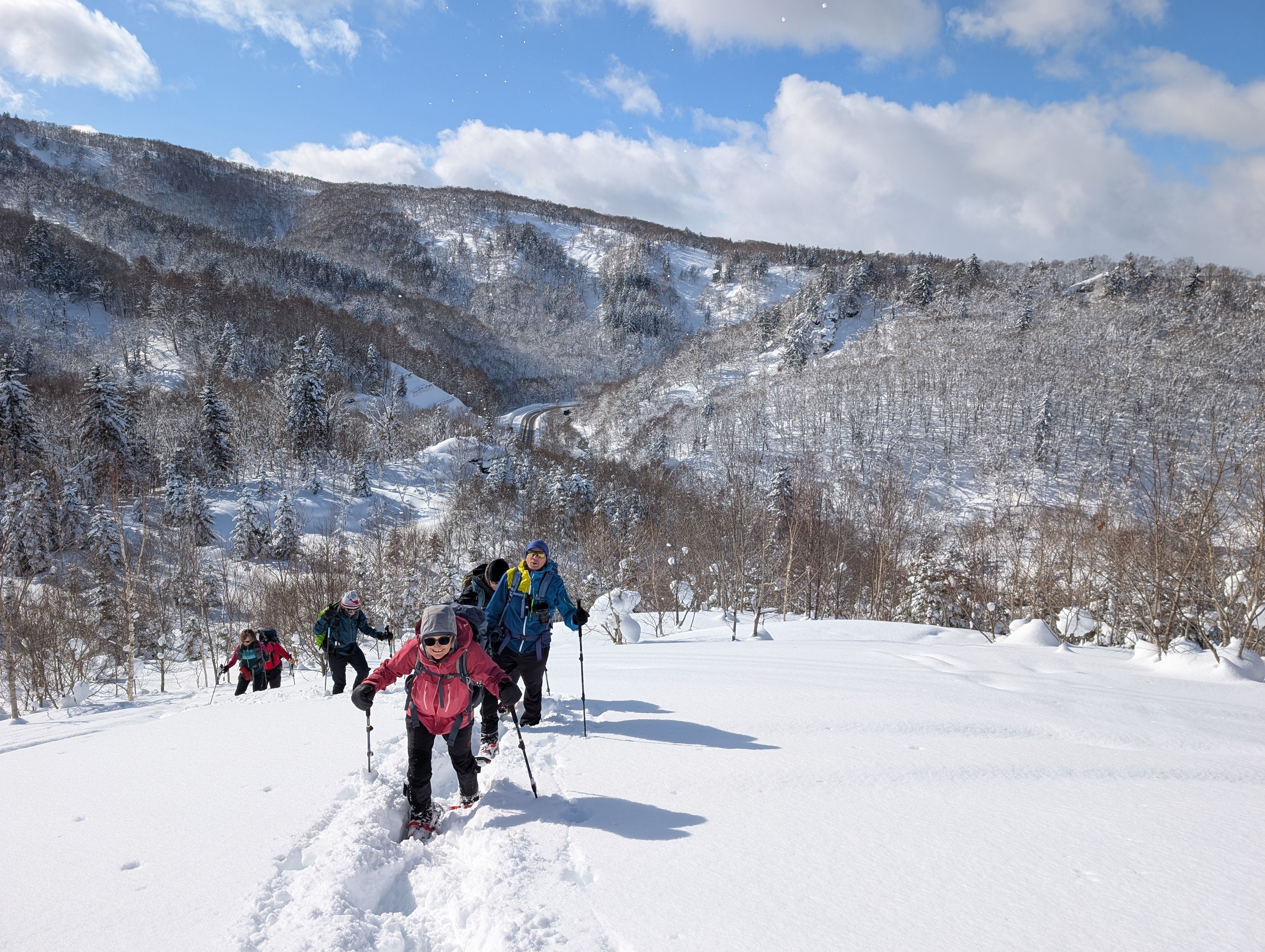 A line of hikers walk up through the snow on Mt. Kokimobetsu It is a sunny day and there are blue skies overhead.