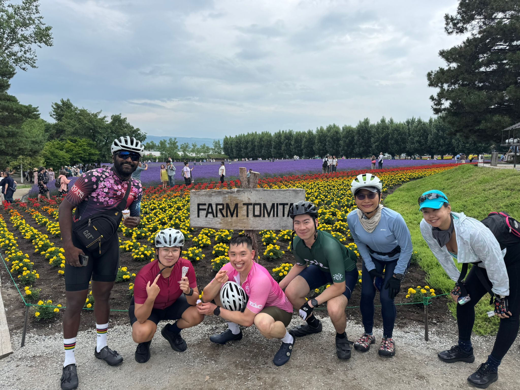 A group of cyclists smile in front of a sign reading "Farm Tomita". A flower meadow full of tourists is visible behind them.
