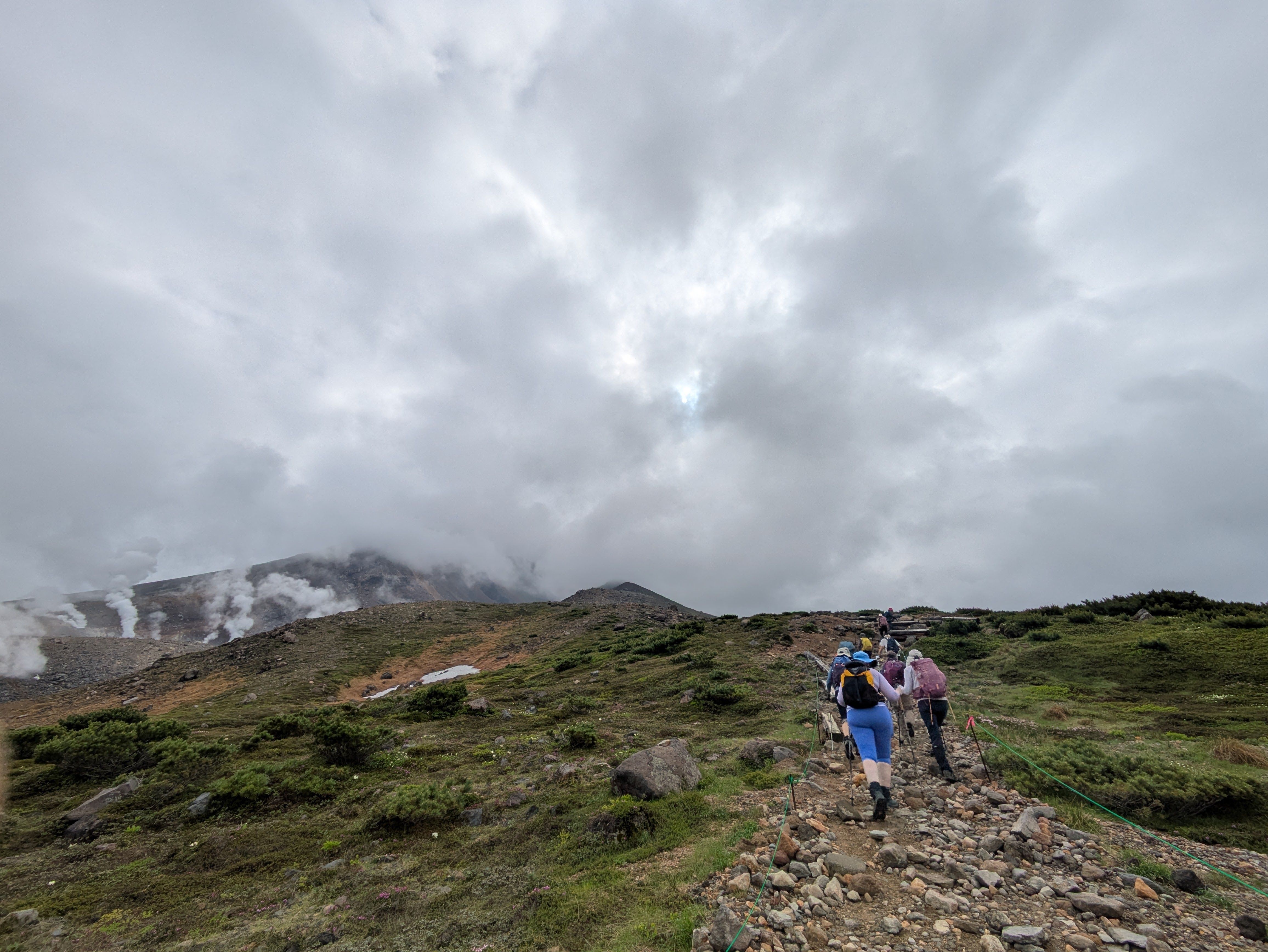 A group of hikers walks up the route to the summit of Mt. Asahidake. They pass by a crater from which steam vents out of volcanic fumaroles.
