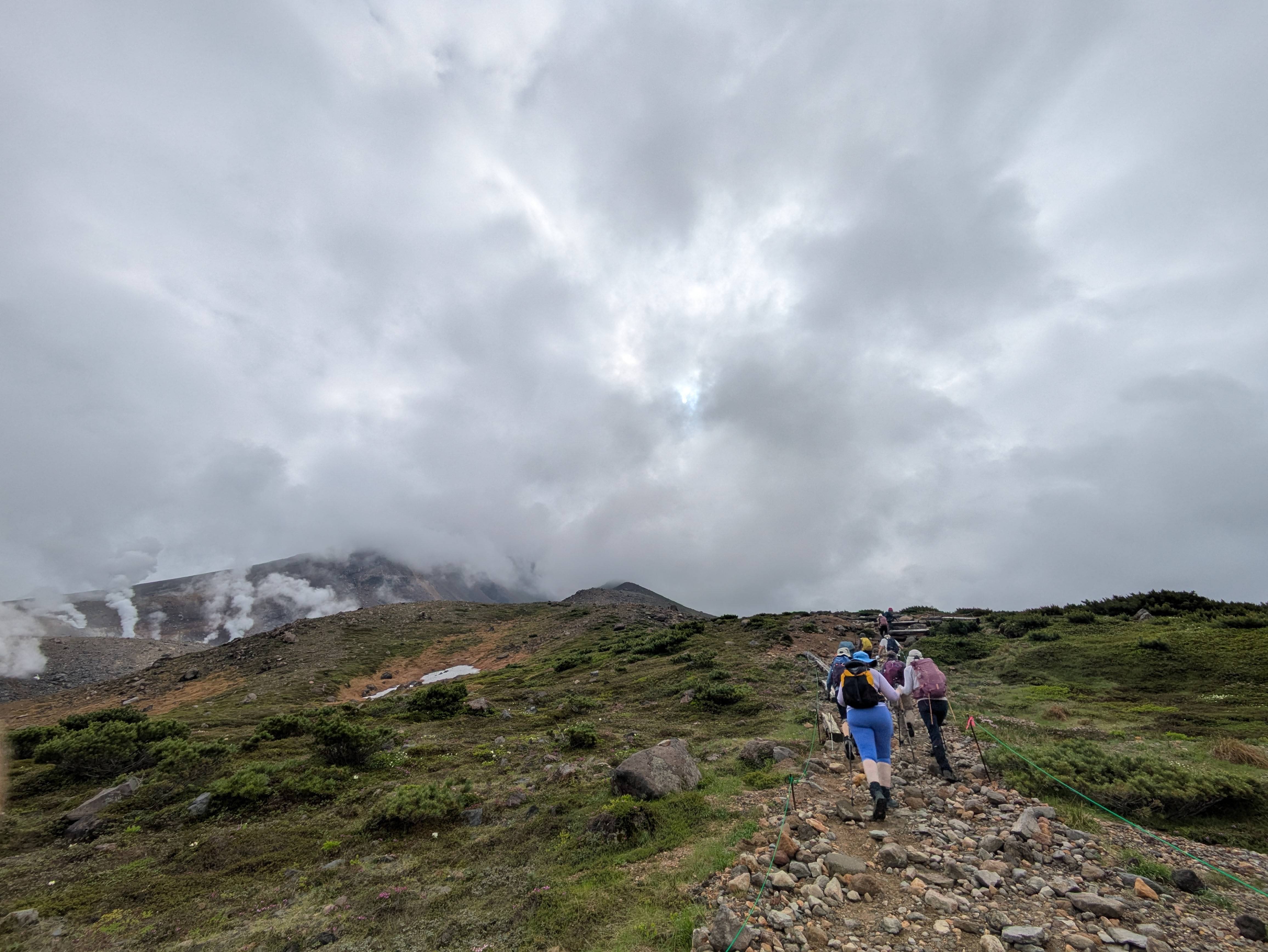 A group of hikers walks up the route to the summit of Mt. Asahidake. They pass by a crater from which steam vents out of volcanic fumaroles.