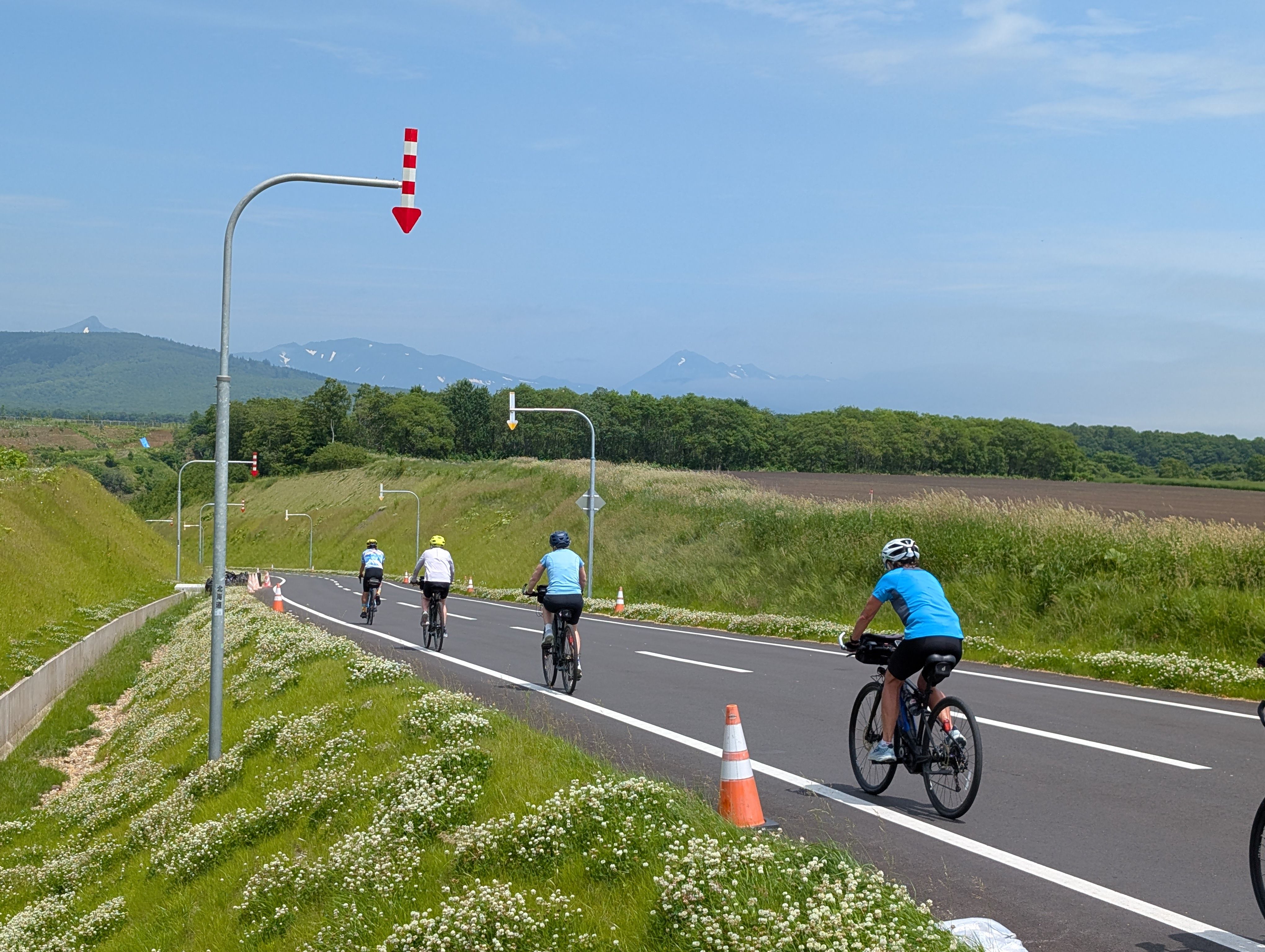 A group of cyclists descend on a road. It is a sunny day. Flowers grow in patches at either side of the road. In the distance, the mountains of the Shiretoko Peninsula are visible. Snow patches remain on the mountains.