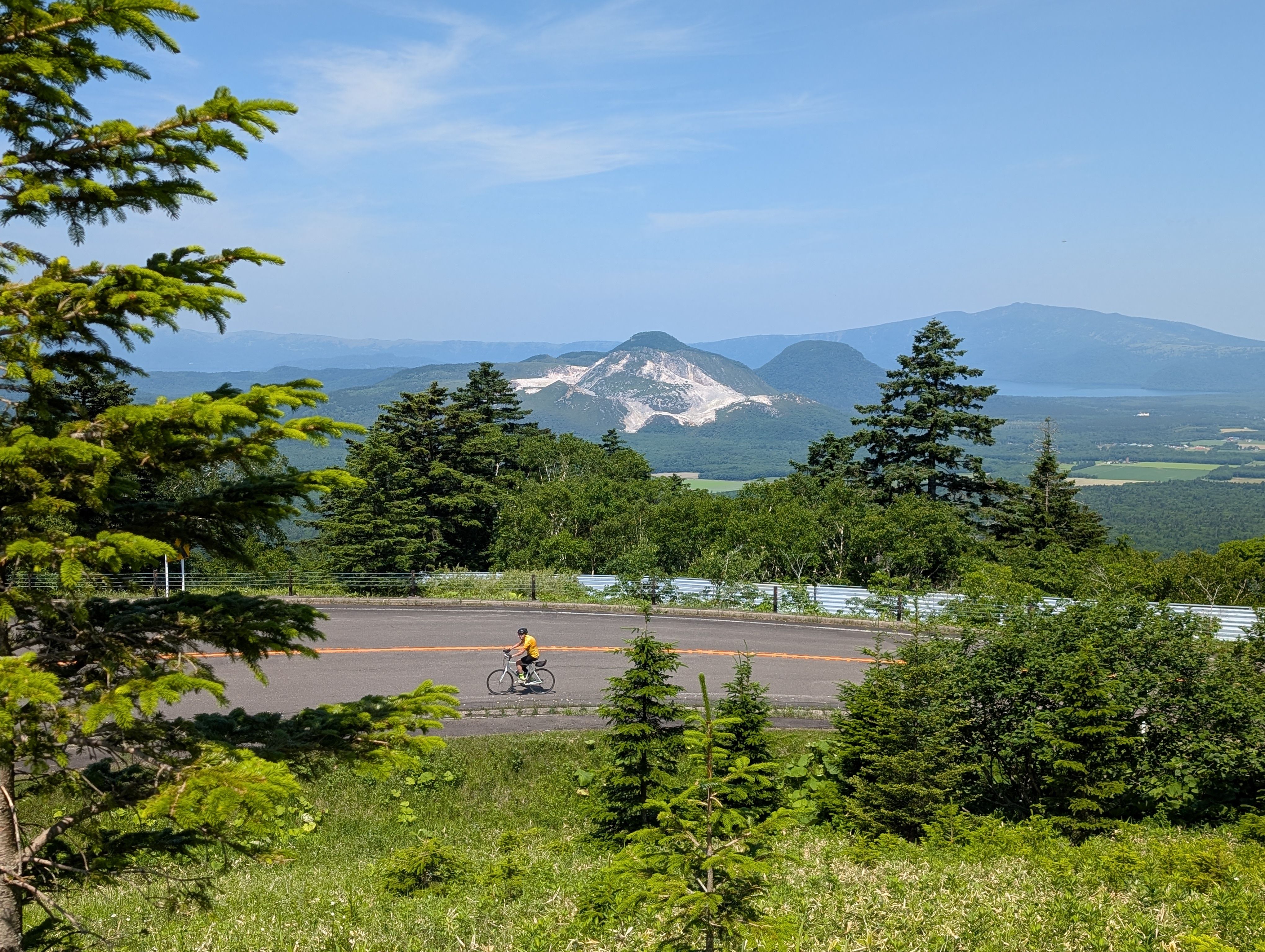 A cyclist ascends a mountain road. In the background. Mt. Io, an active volcano, is visible. It is a bright and sunny day.