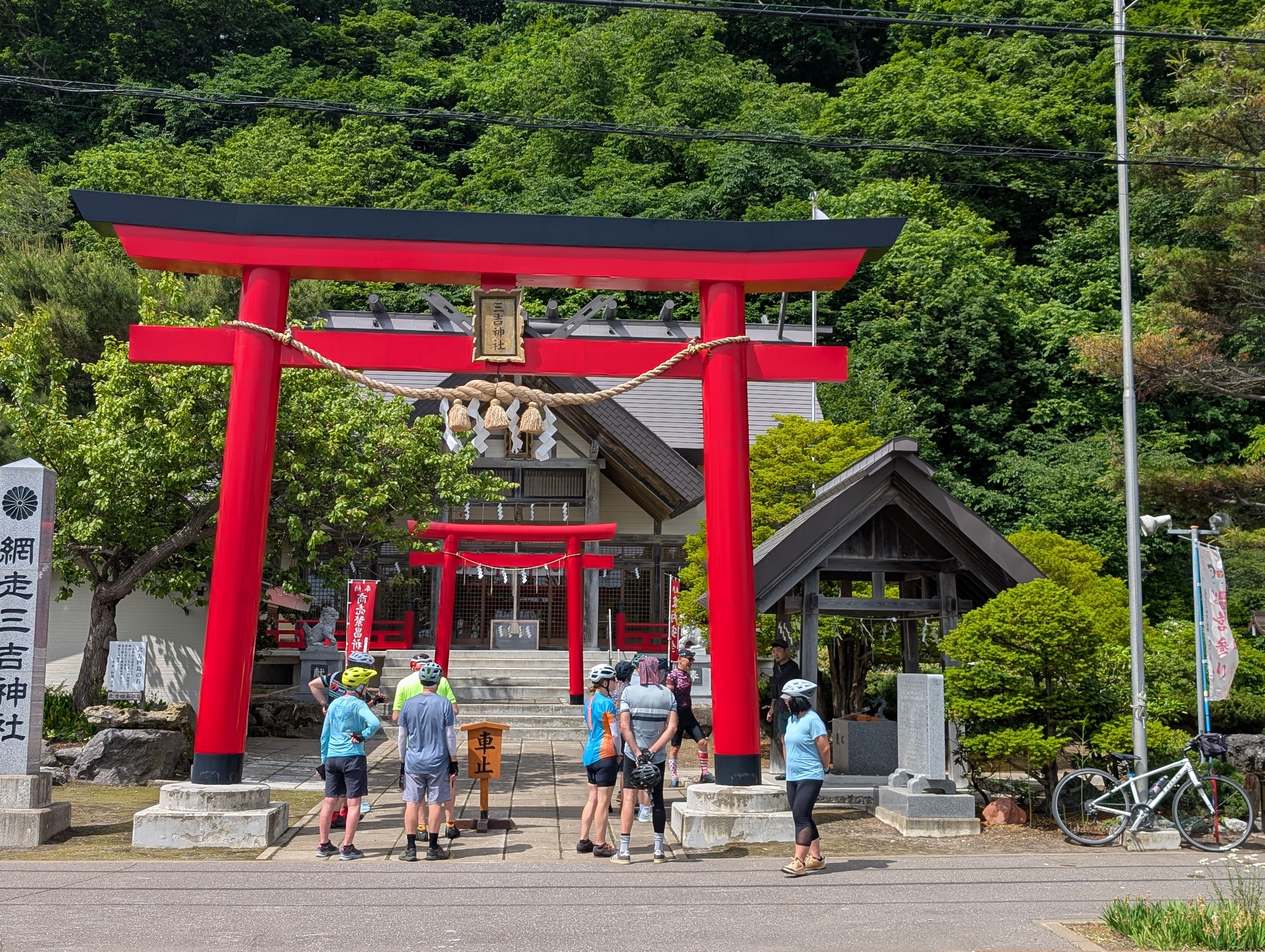 A group of cyclists stand in front of a large, red Torii gate at the entrance to a Japanese shrine.