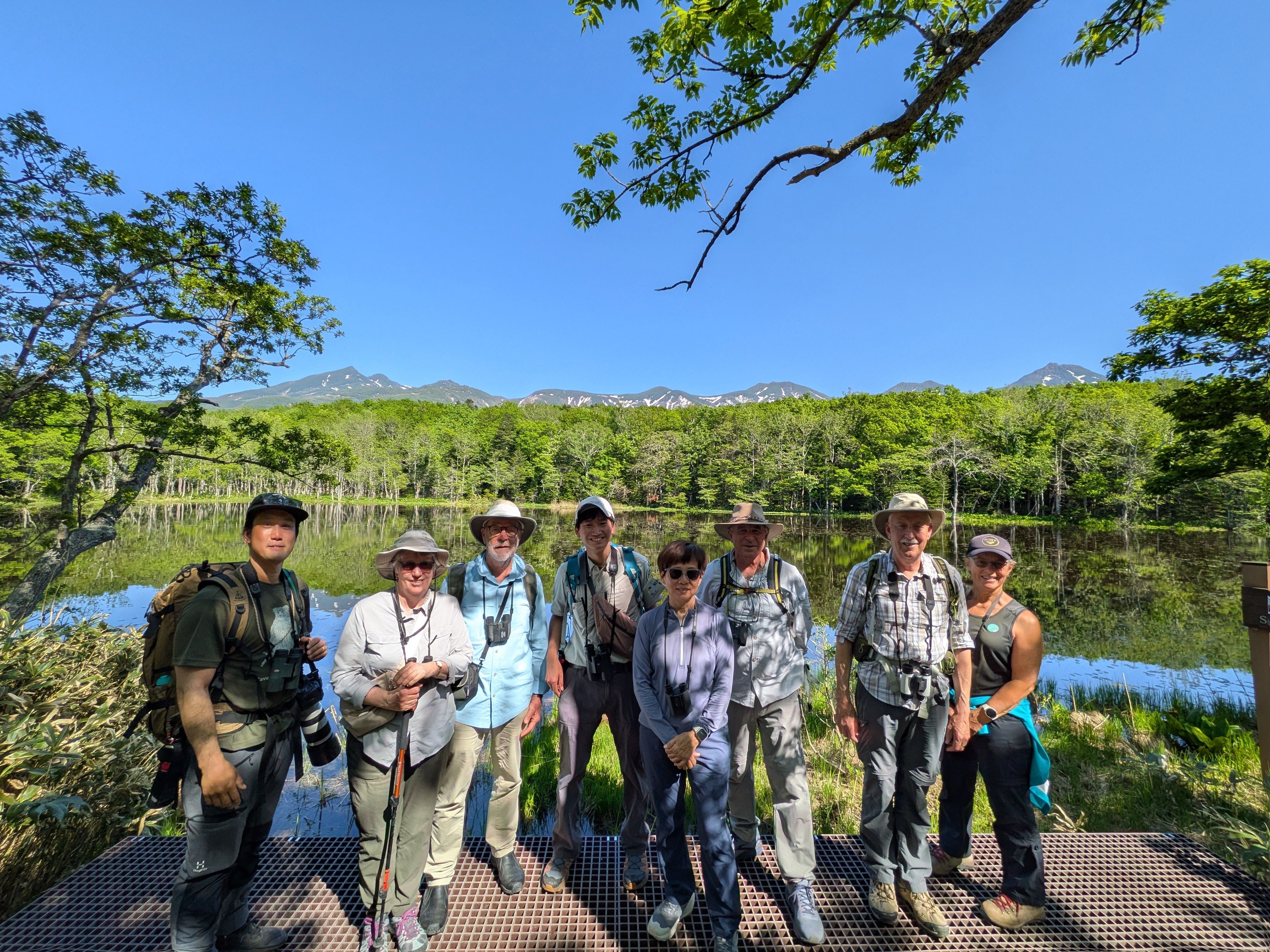 A group of hikers stand in a line in front of one of the Shiretoko Five Lakes in Shiretoko Peninsula, Hokkaido. They are smiling at the camera. It is a cloudless day and the mountain range behind them is reflected in the lake like a mirror.