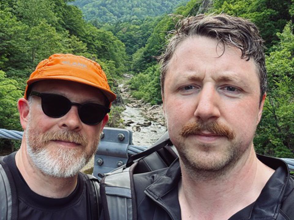 Two men, one with a beard and one with a moustache, looking at the camera while taking a selfie. They are on a bridge and a river flanked with steep forested slopes, making the scene vibrant and green.