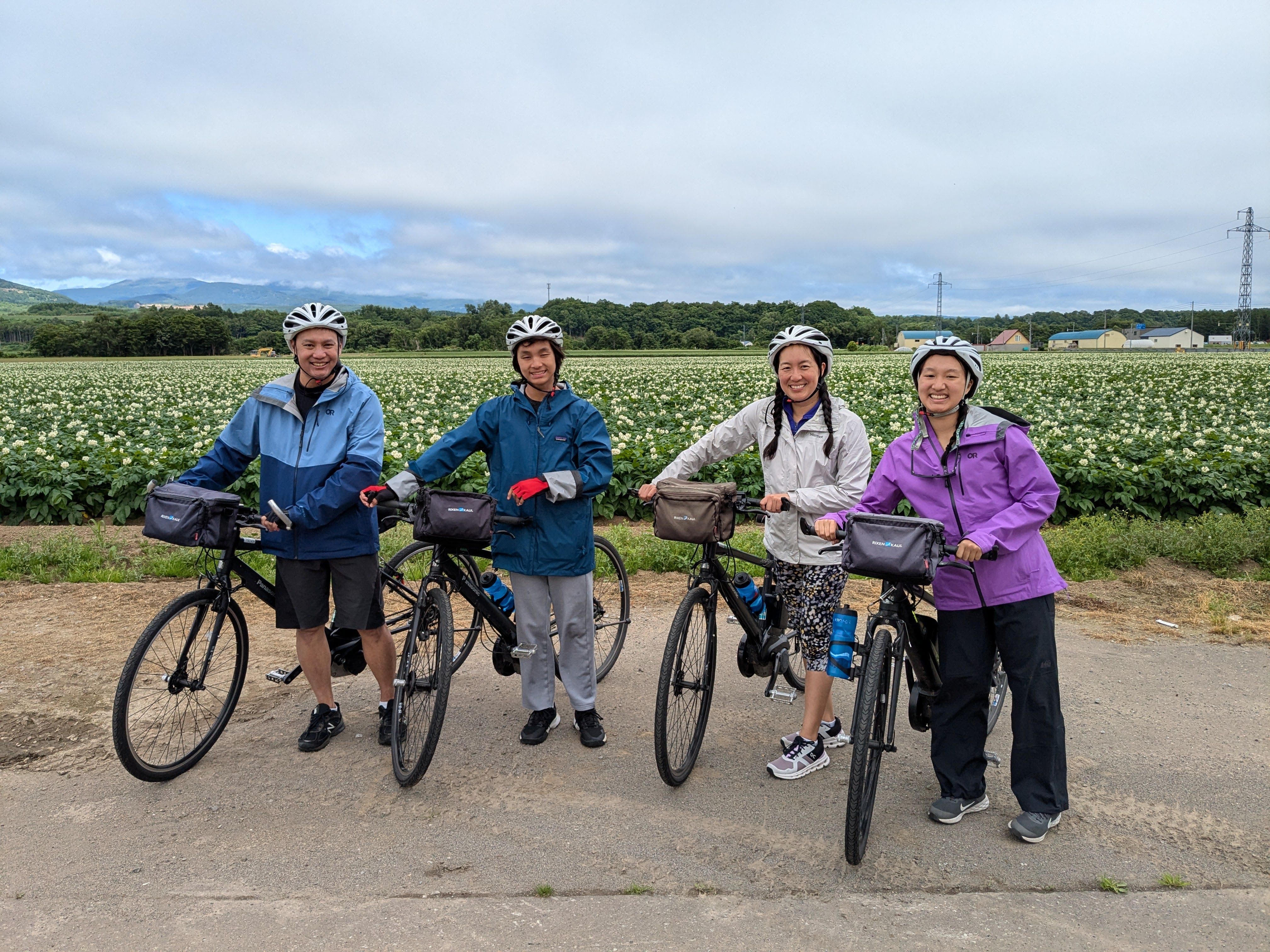 A group of four cyclists pose and smile in front of a field of potatoes in Hokkaido. The potato flowers are in bloom, indicating it is spring. It is a cloudy day.