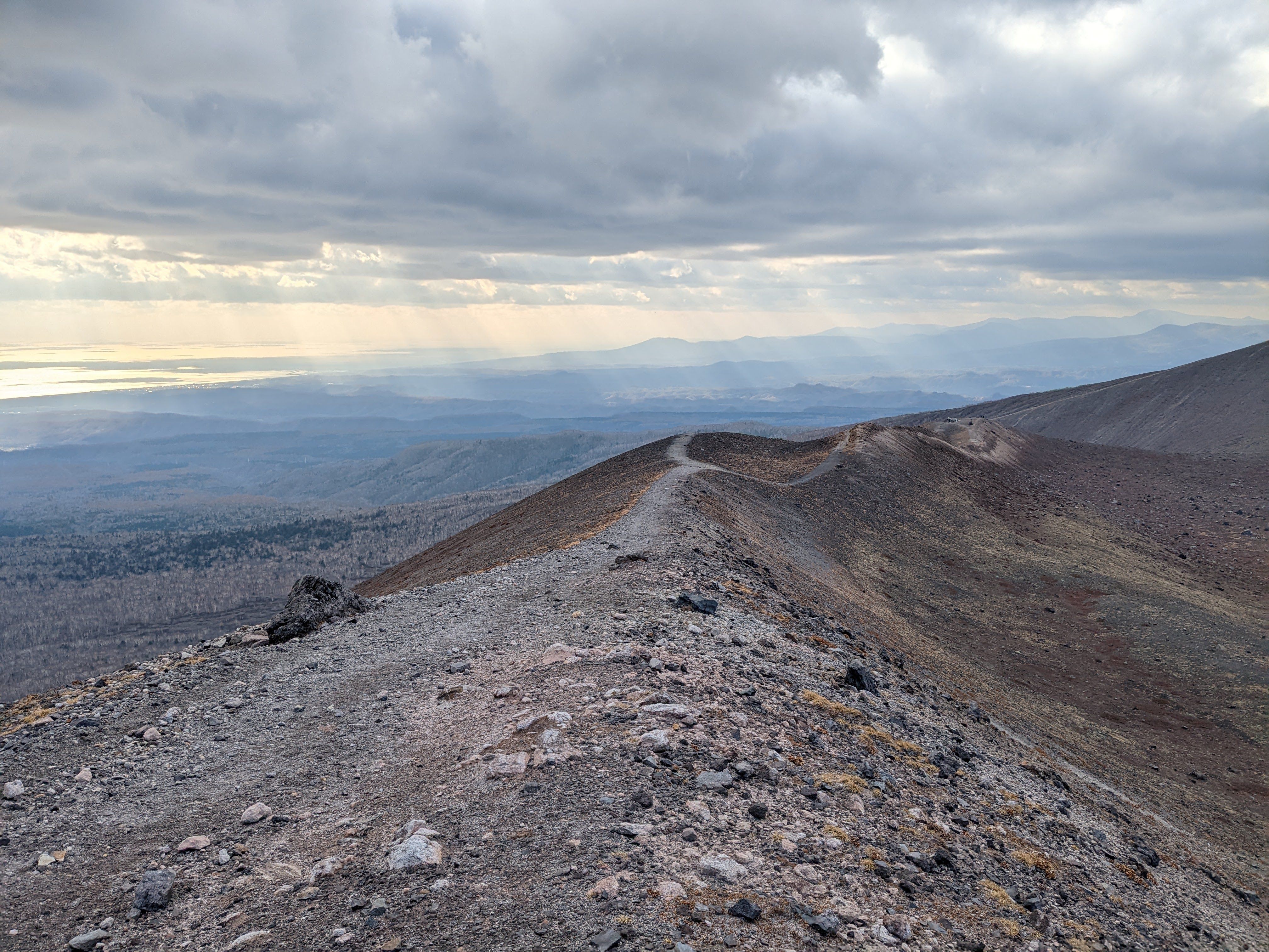 A rocky trail runs away to into the distance along an exposed ridge line flanked by a barren volcanic landscape. In the distance, rays of sun can be seen streaking out of clouds and falling on the low lying areas. The ocean is seen catching the sun on the left.