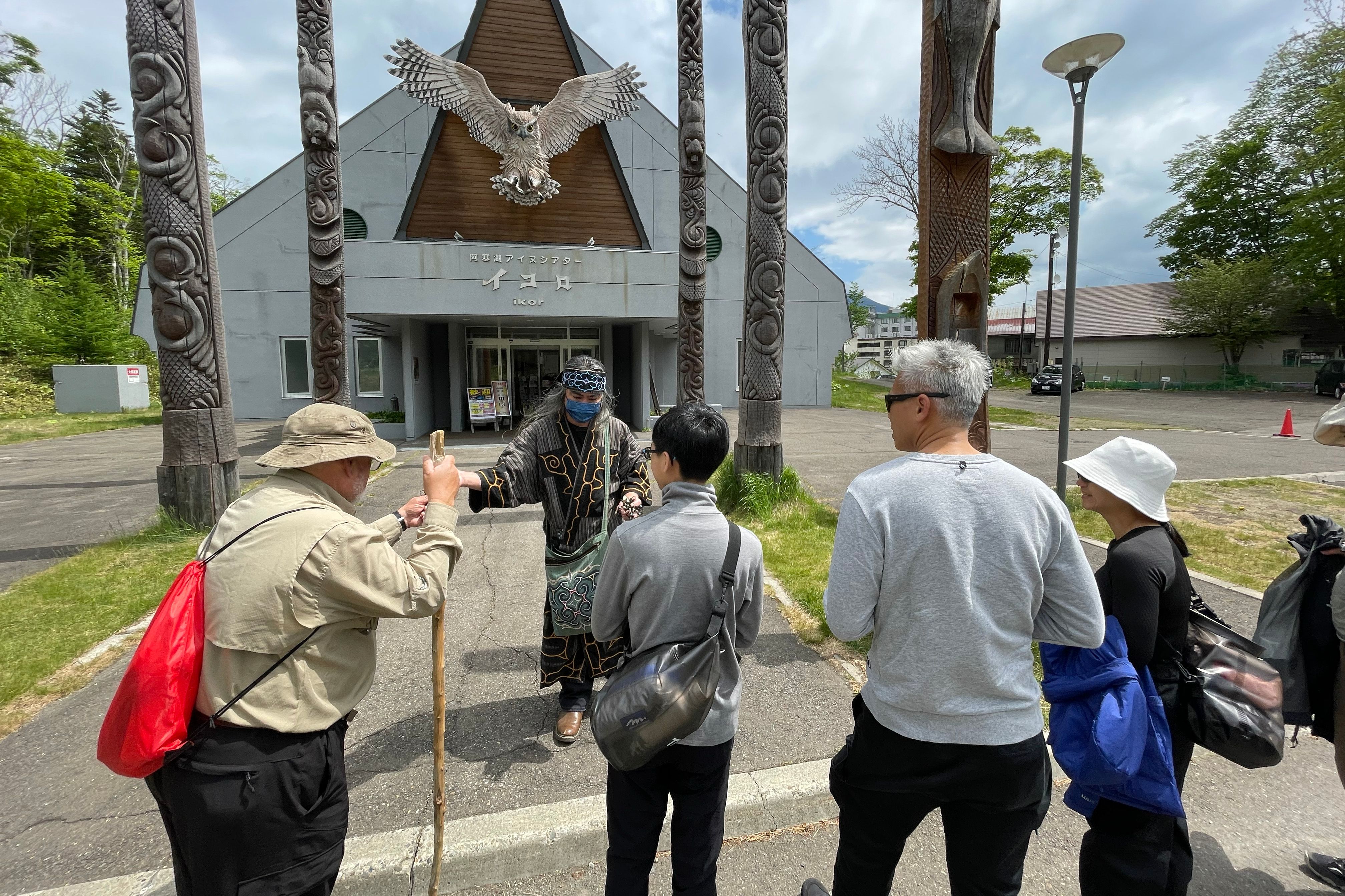 A group of tourists are greeted by an Ainu guide outside a building with a large owl sculpture above the door