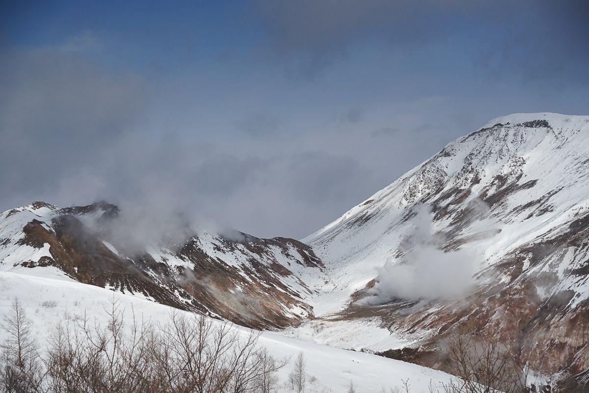 White steam rises from the rugged, volcanic crater of Mt. Usu in Toya-Shikotsu National Park. The winter landscape features steep slopes covered in white snow, revealing patches of brown rock, set against a cloudy blue sky.