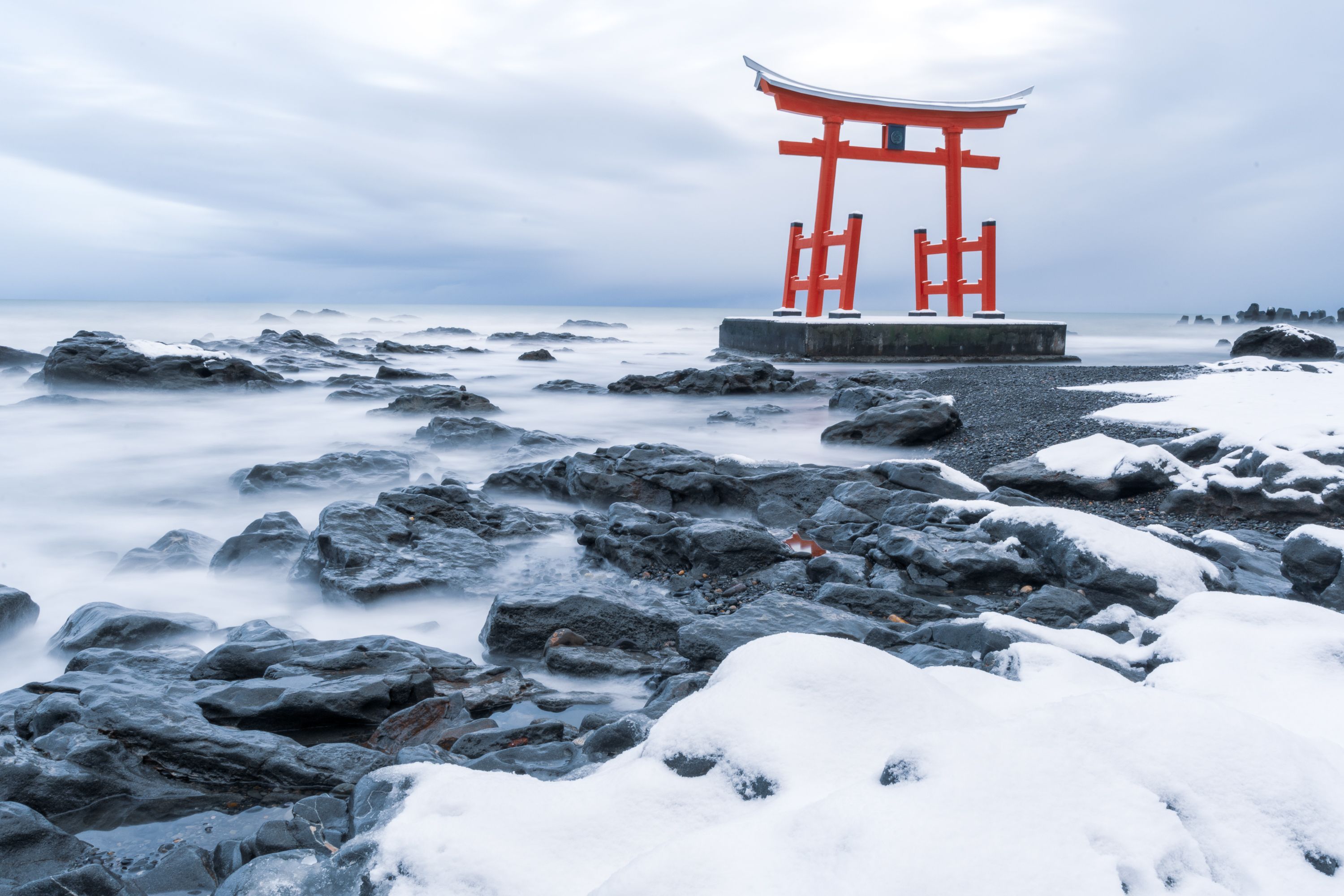 A long-exposure photo of a shrine gate stood on a rocky shoreline. The waves have been taken over a long period so that they appear like mist in the photograph.