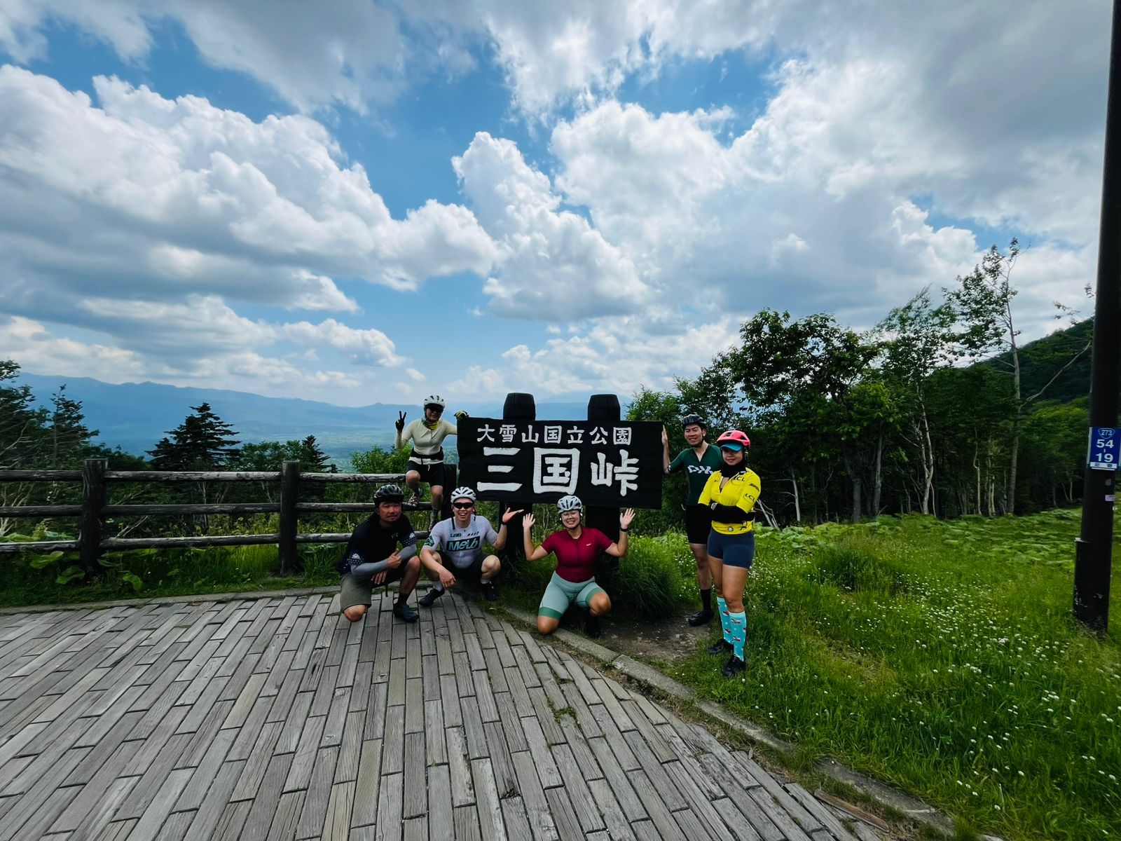 A group of cyclists pose in front of a sign reading "Daisetsuzan National Park - Mikuni Pass" in Japanese. They are all smiling at the camera. It is a partially-cloudy day.
