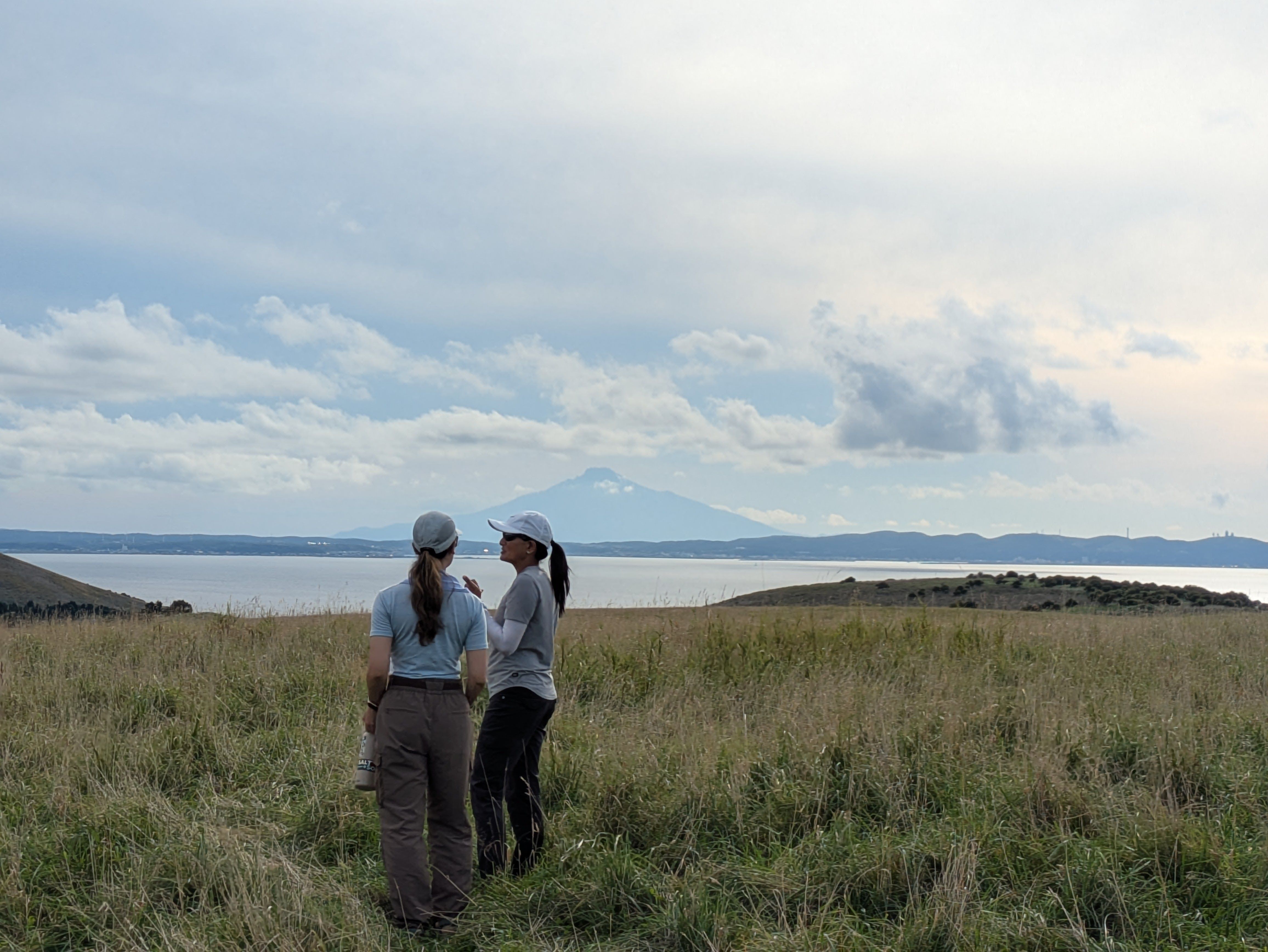 Two women stand in grassland just off the White Shell Path in Wakkanai, Hokkaido. The ocean is visible beyond and, far off in the distance, the conical silhouette of Mt. Rishiri is visible.