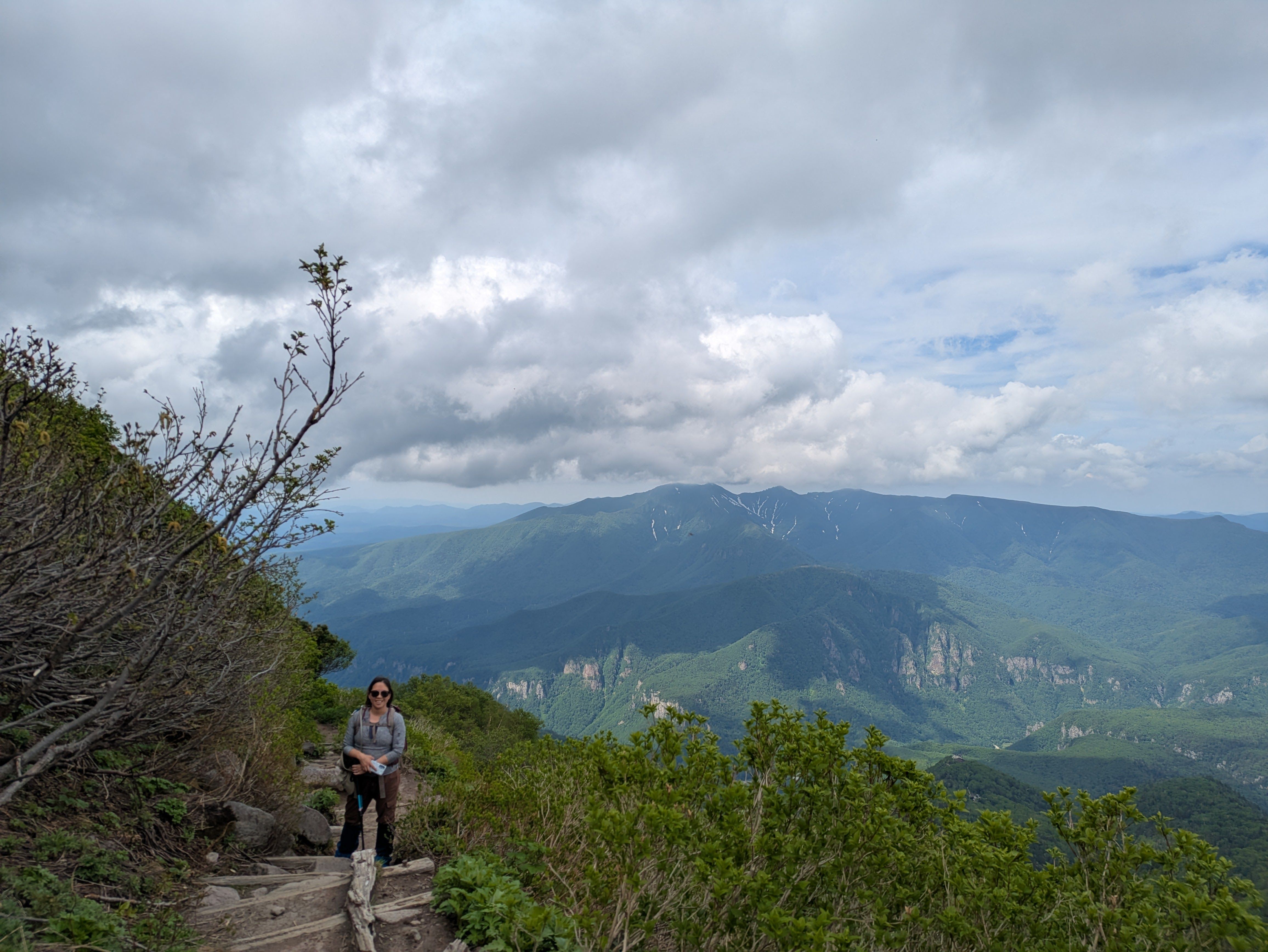 A woman on a hiking trail on Mt. Kurodake, Hokkaido, smiles at the camera. Green mountainous valleys are visible in the distance behind her.