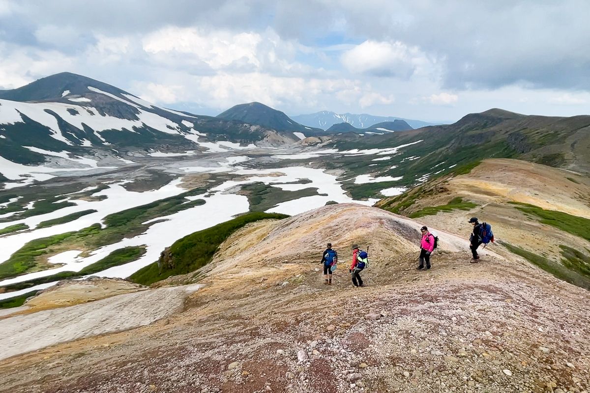 Four hikers gaze upon Daisetsuzan's Ohachidaira in summer, where large patches of snow remain.