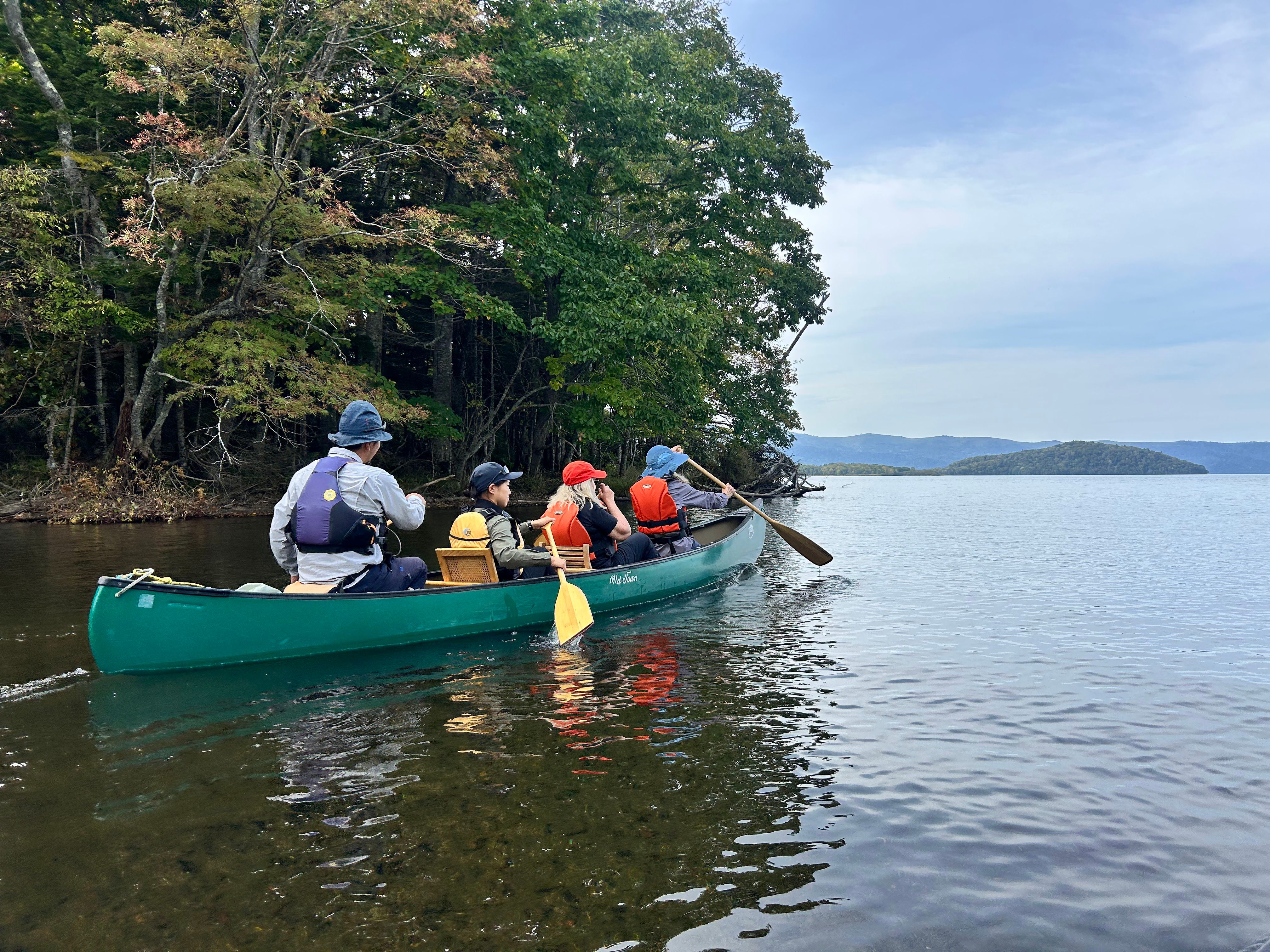 Four people paddle a canoe out onto the open waters of Lake Kussharo. On the far bank is a forest. Mountains are visible on the far side of the lake. The water is deep, still and clear.