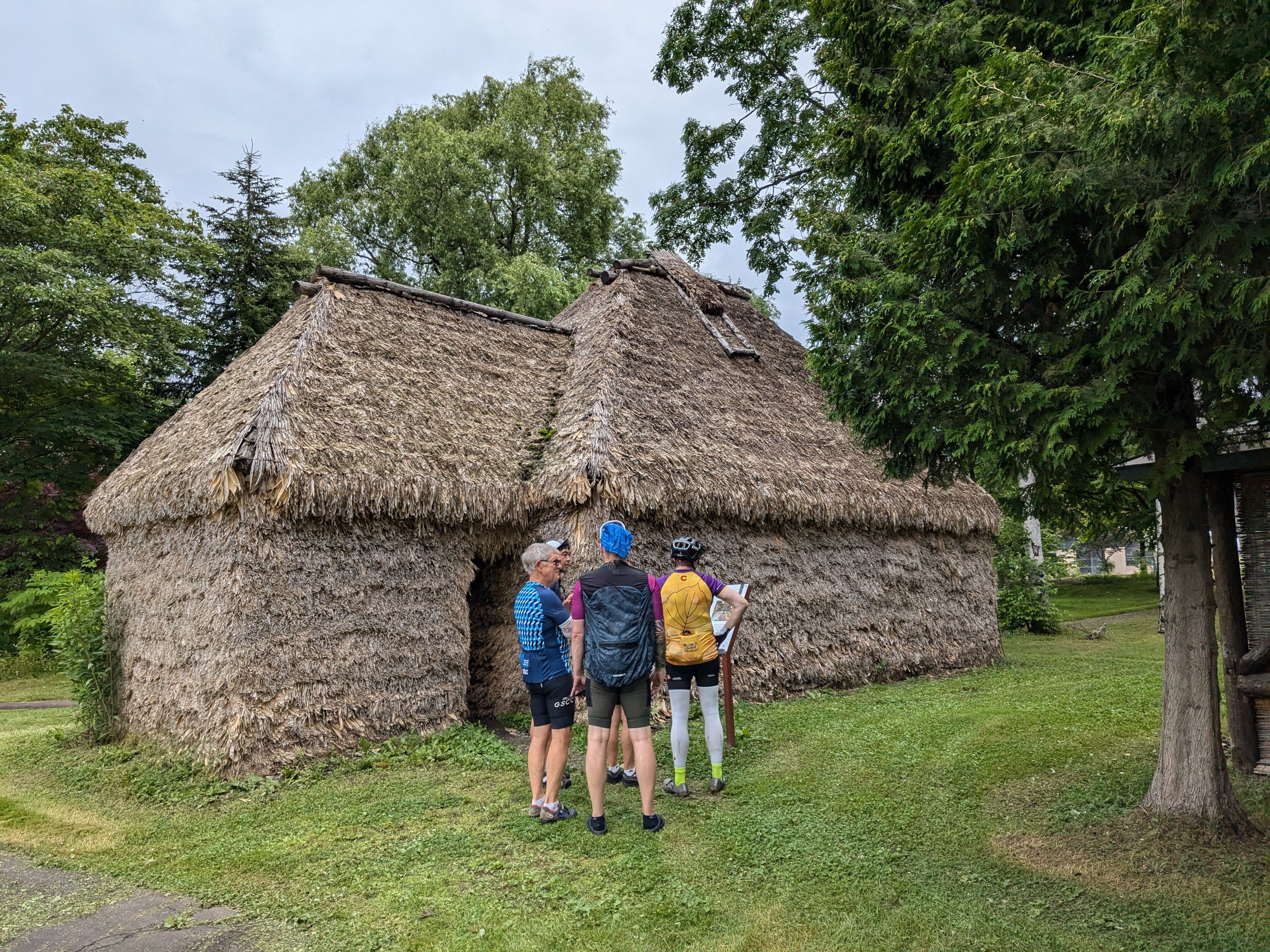 A group of cyclists stand outside a cise, a traditional Ainu dwelling made from reeds and bamboo leaves.