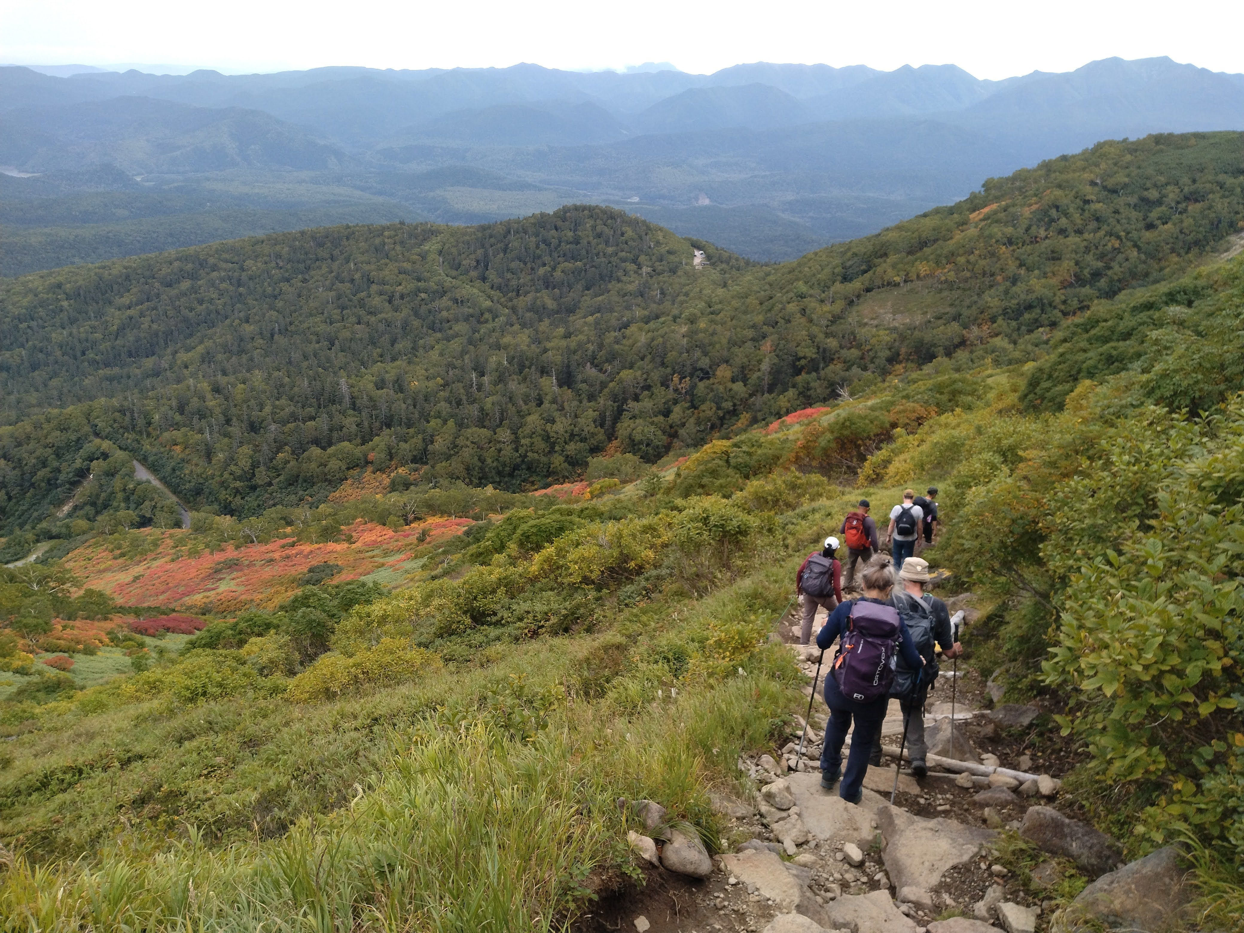 A group of hikers descends Mt. Aka in Daisetsuzan National Park, Hokkaido. In the mountain valley below, reddening autumn leaves are visible.