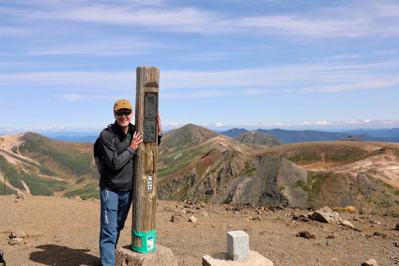 A happy hiker, Adrian from the UK, smiles while standing at the summit of Mt. Asahidake in Hokkaido. He is holding onto a wooden post with Japanese writing, with a vast, mountainous landscape stretching out behind him under a blue sky.