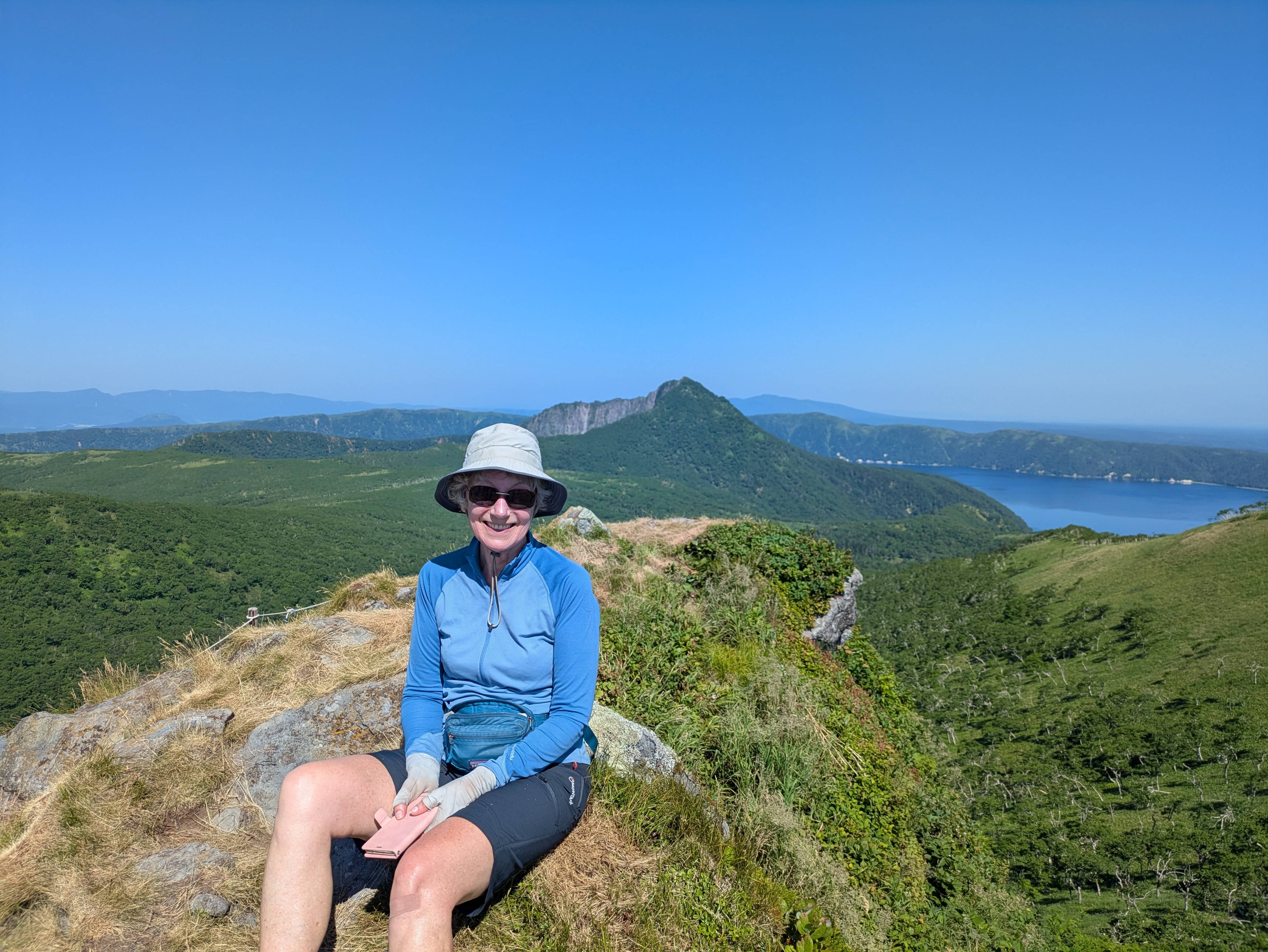 A woman smiles at the camera at the summit of Mt. Risuke in Hokkaido, Japan. Behind her is Lake Mashu. It is a sunny day with pure blue skies overhead.