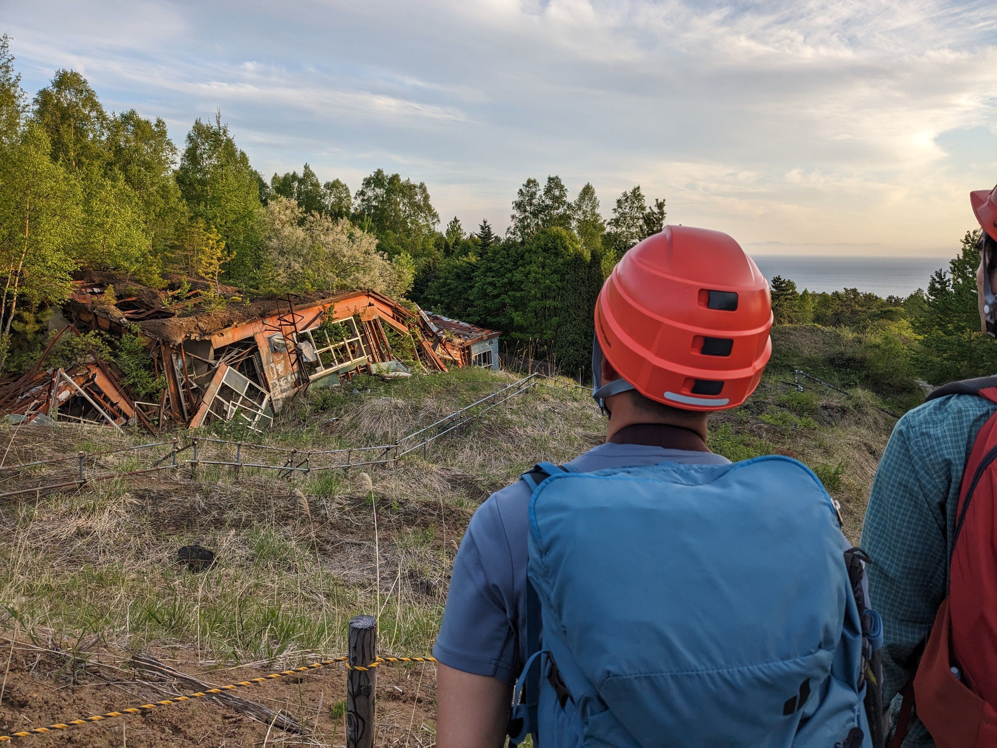 Two individuals wearing hiking gear and helmets stand with their backs to the camera, looking out at the aftermath of the Mt. Usu eruption. In the mid-ground, a broken, buckled, reddish-brown house lies in ruins amidst dry, overgrown grass and lush green trees. In the distance, a calm blue sea meets the horizon under a cloudy sky.
