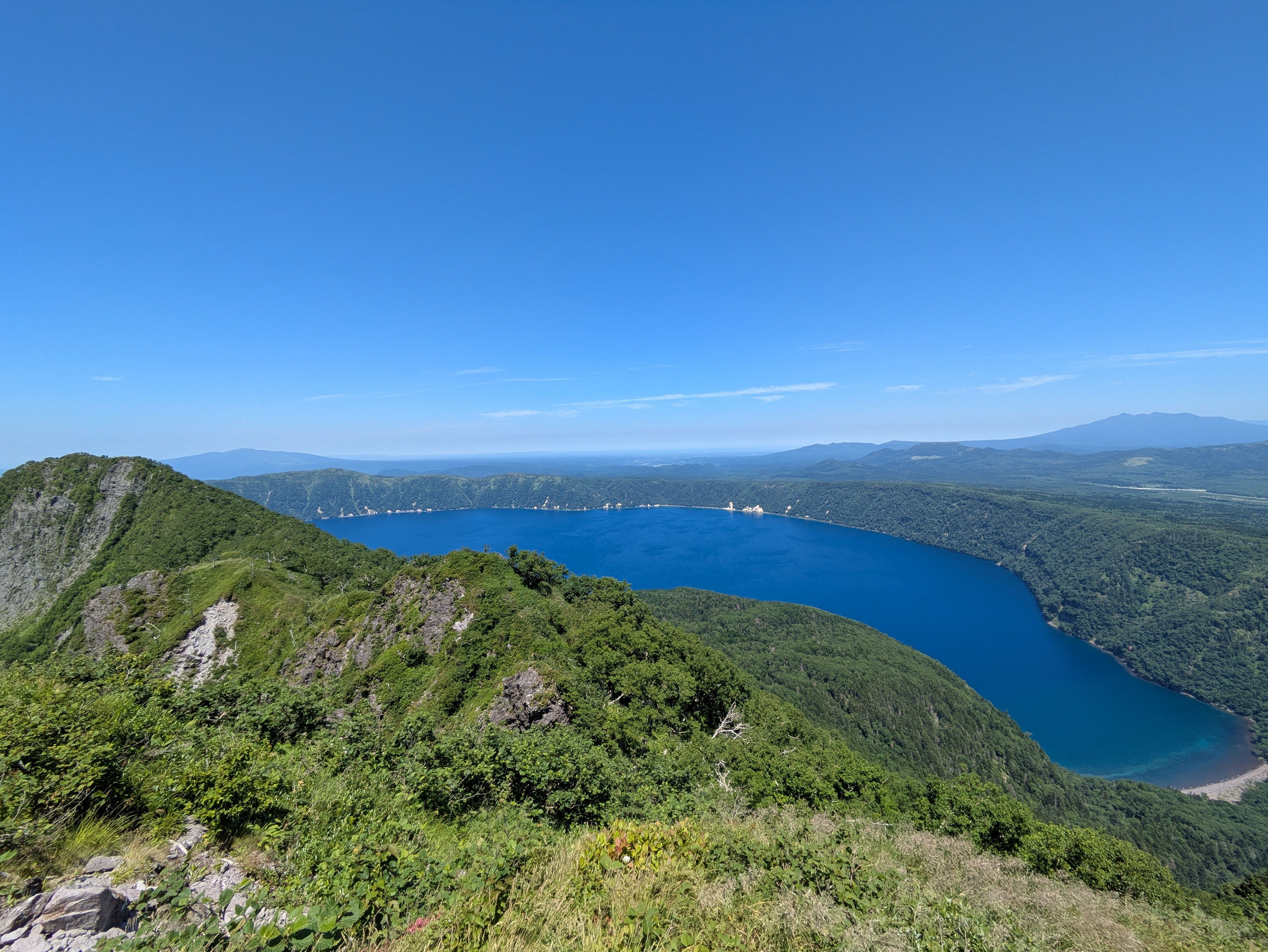 A view of Lake Mashu from the summit of Mt. Risuke in Hokkaido. It's a sunny day and the surface of the lake is a beautiful, deep blue.