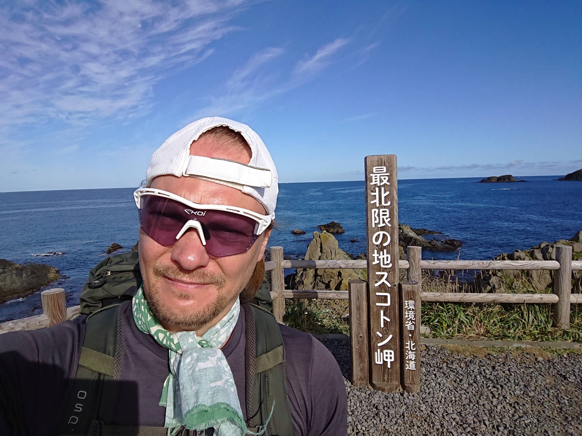 An image of a man in sunglasses and a backwards baseball cap taking a selfie in front of a guidepost and the ocean. The guidepost reads "Cape Sukuton, Northernmost Point in Japan".
