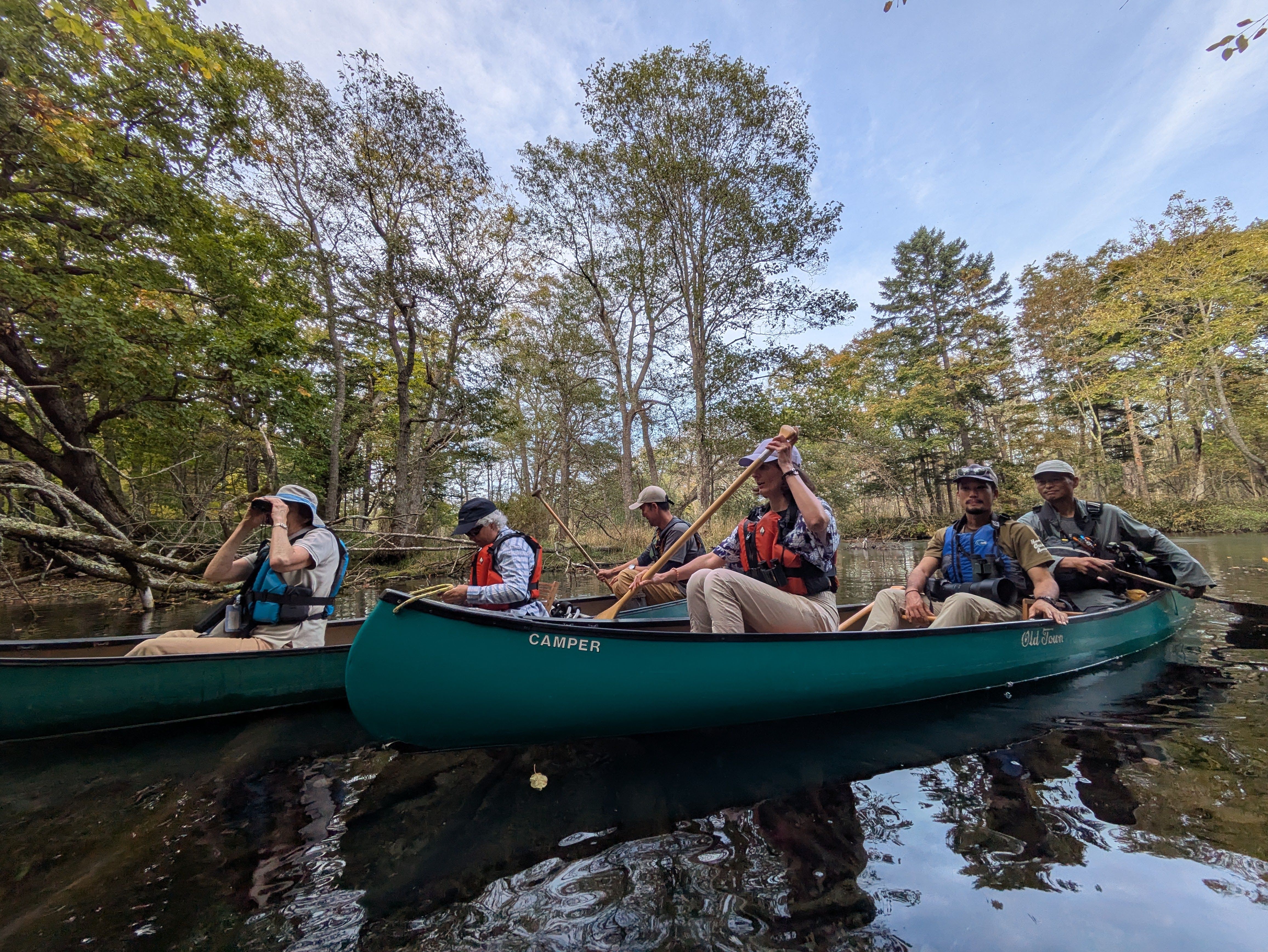 Two canoes, three people seated in each, cruise alongside each other on the Kushiro River. There is forest around them and it is a beautiful day with blue skies above.