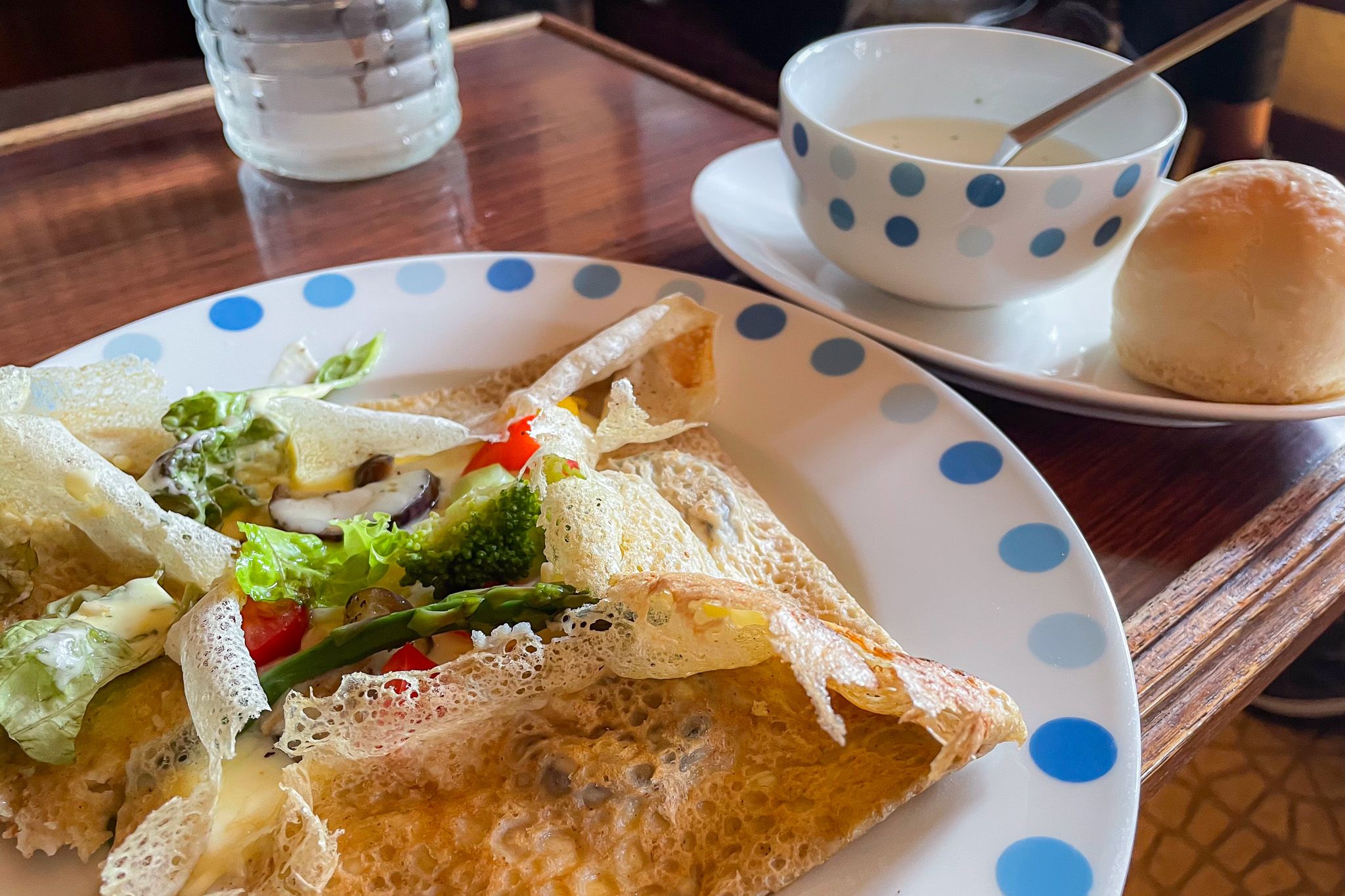 A galette (crepe dish filled with egg, cheese and vegetables) on a plate on a wooden table. Behind it is a small bowl of soup with a bread roll.