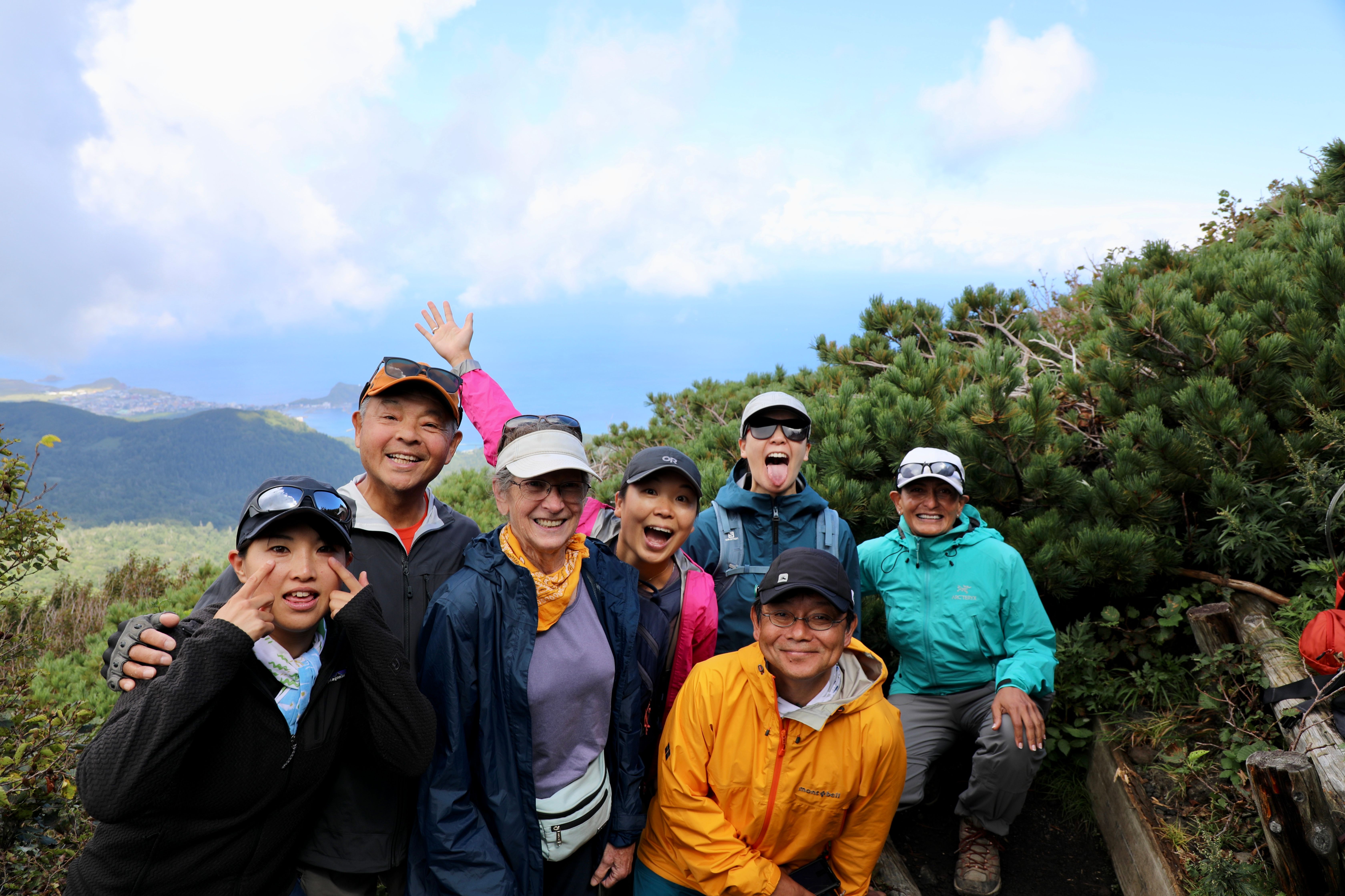 A group of hikers smile at the camera on a mountain trail, scenery just coming out of the clouds behind them. Some of them pull funny faces at the camera. (Photo Copyright: Samir Patel)